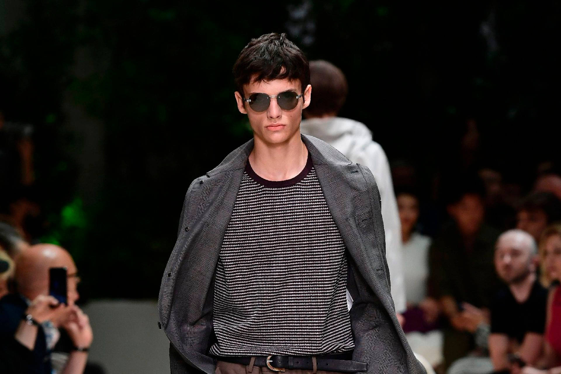 Ferragamo 桌布 壁紙 — 一位男模特兒自信地走在伸展台上，戴著太陽眼鏡、灰色西裝外套和黑白圖案襯衫，背景是模糊的觀眾。