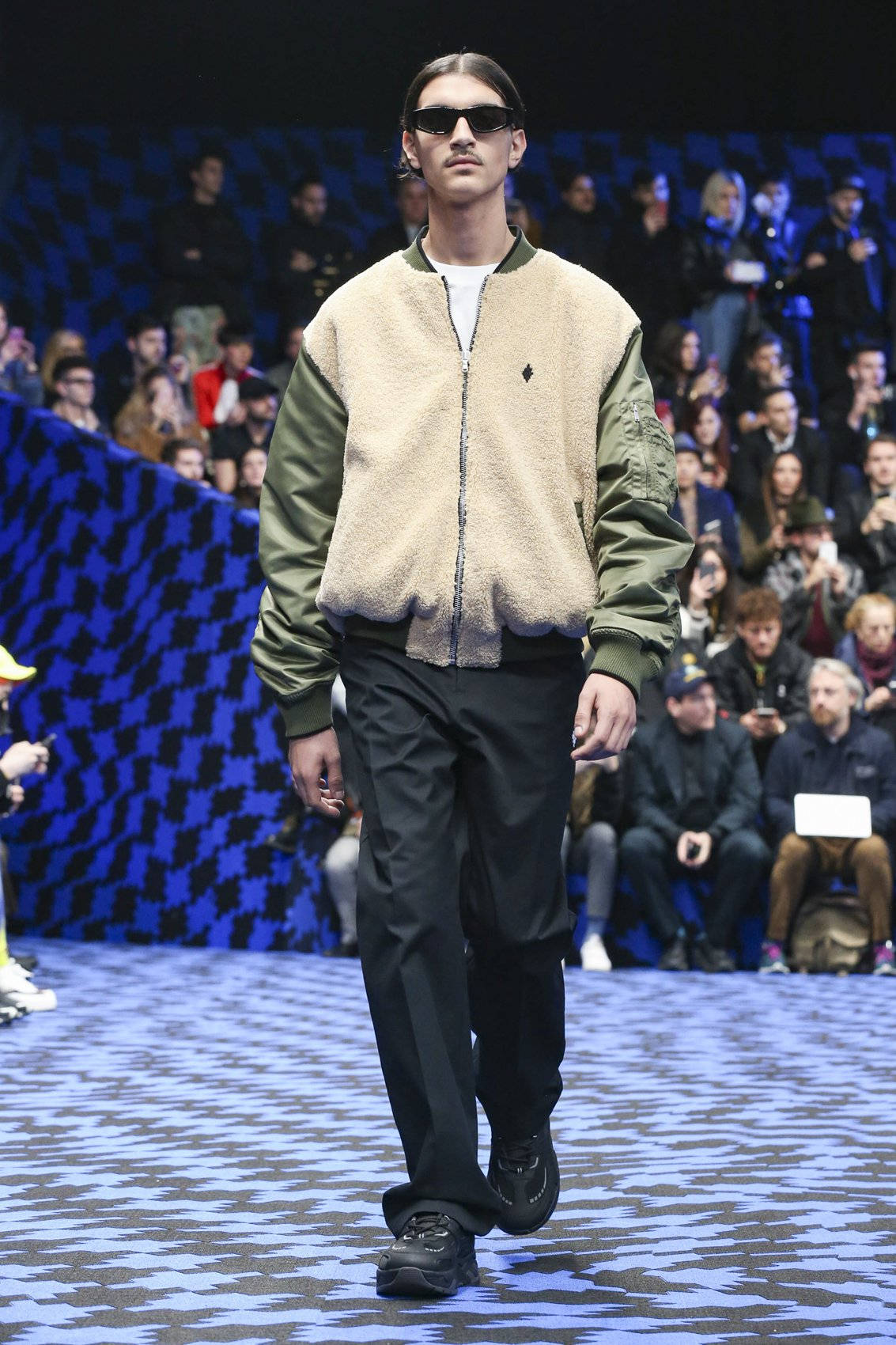 Fashion Model 桌布 壁紙 — 一位男模特兒自信地走在伸展台上，身穿米色和橄欖色飛行員夾克、黑色褲子和太陽眼鏡，在明亮的舞台燈光下。