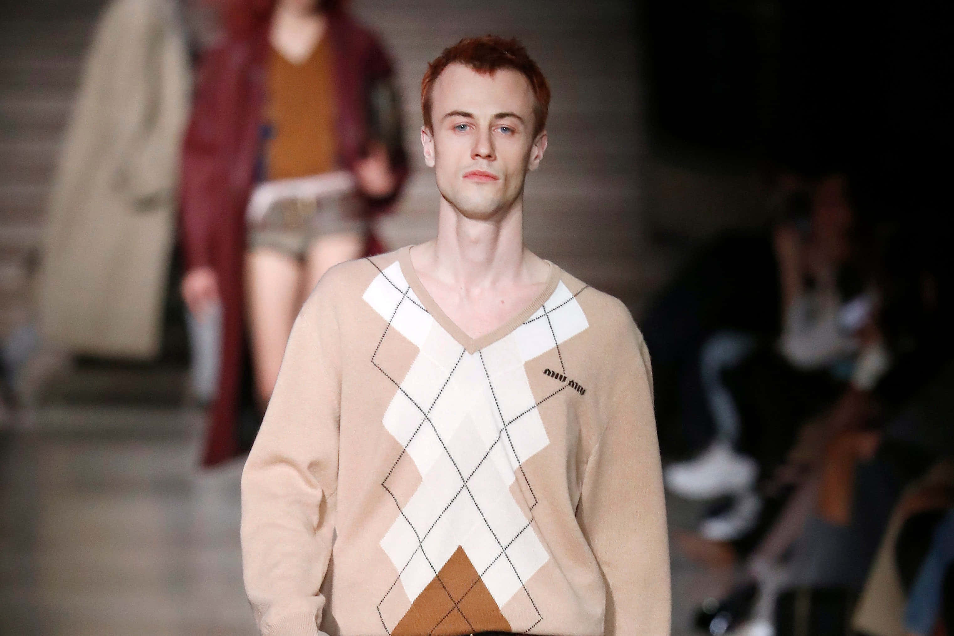 Mens Fashion 桌布 壁紙 — 一位男模特兒自信地走在燈光昏暗的伸展台上，身穿米色菱形花紋毛衣，帶有棕色和白色的幾何圖案。