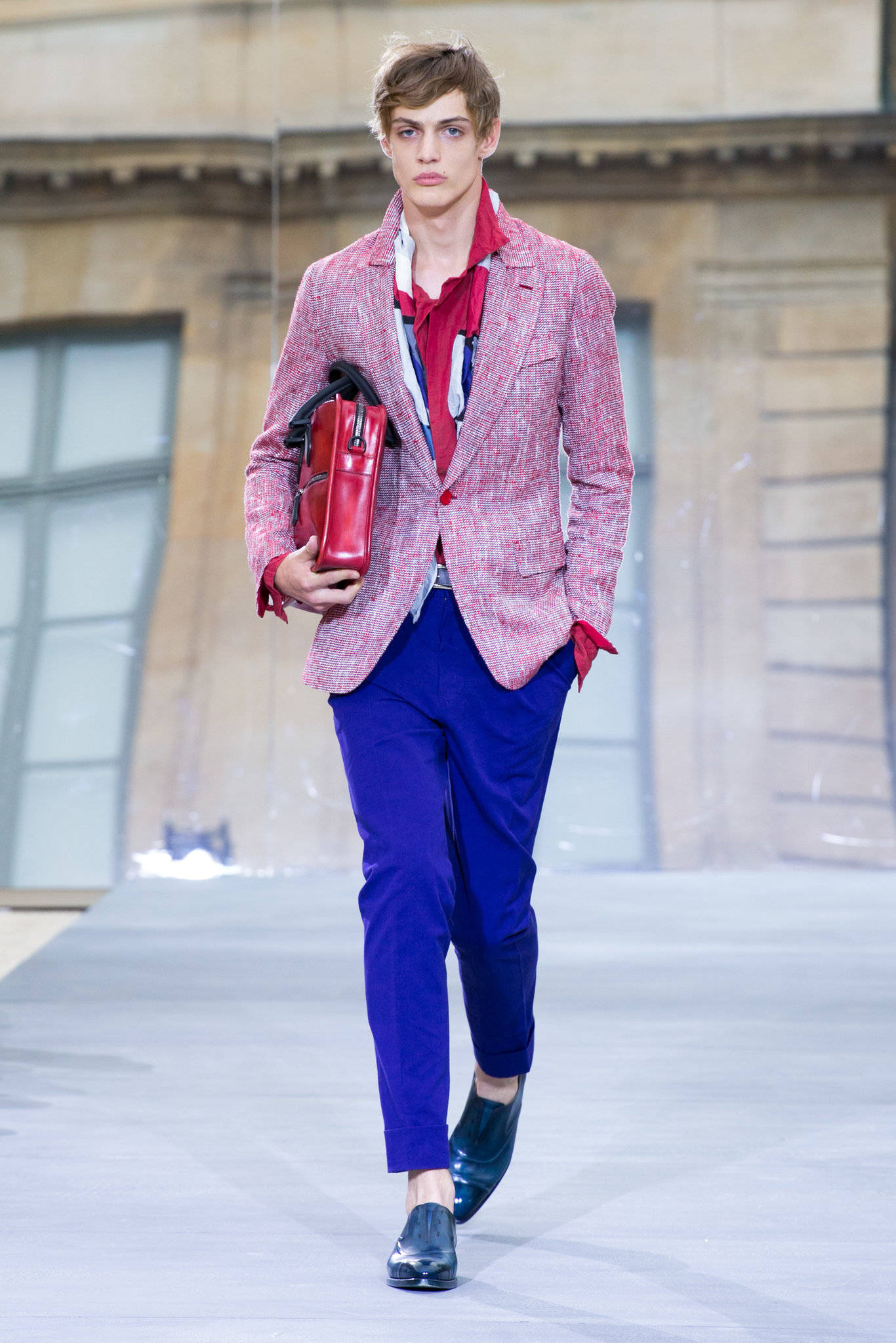 Runway 桌布 壁紙 — 一位男模特兒自信地走過伸展台，身穿大膽的粉紅色西裝外套、藍色長褲和紅色配飾，背景是經典的石建築。