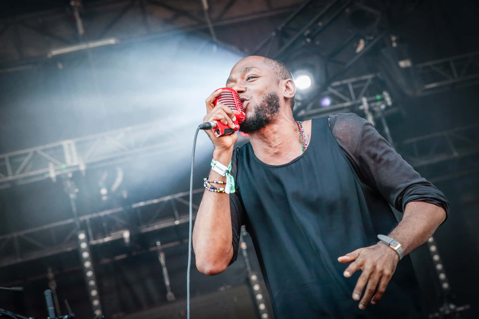 Yasiin Bey 桌布 壁紙 — 一位男歌手在明亮的舞台燈光下，充滿激情地對著復古紅色麥克風演唱，煙霧在他周圍繚繞。