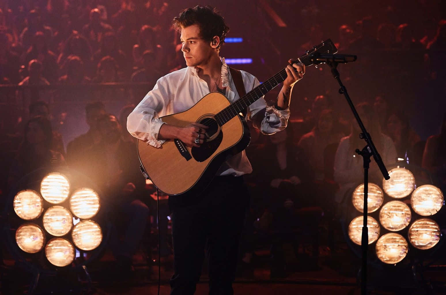 Harry Styles 桌布 壁紙 — 一位男歌手在溫暖的舞台燈光下，在模糊的觀眾面前熱情地彈奏木吉他。