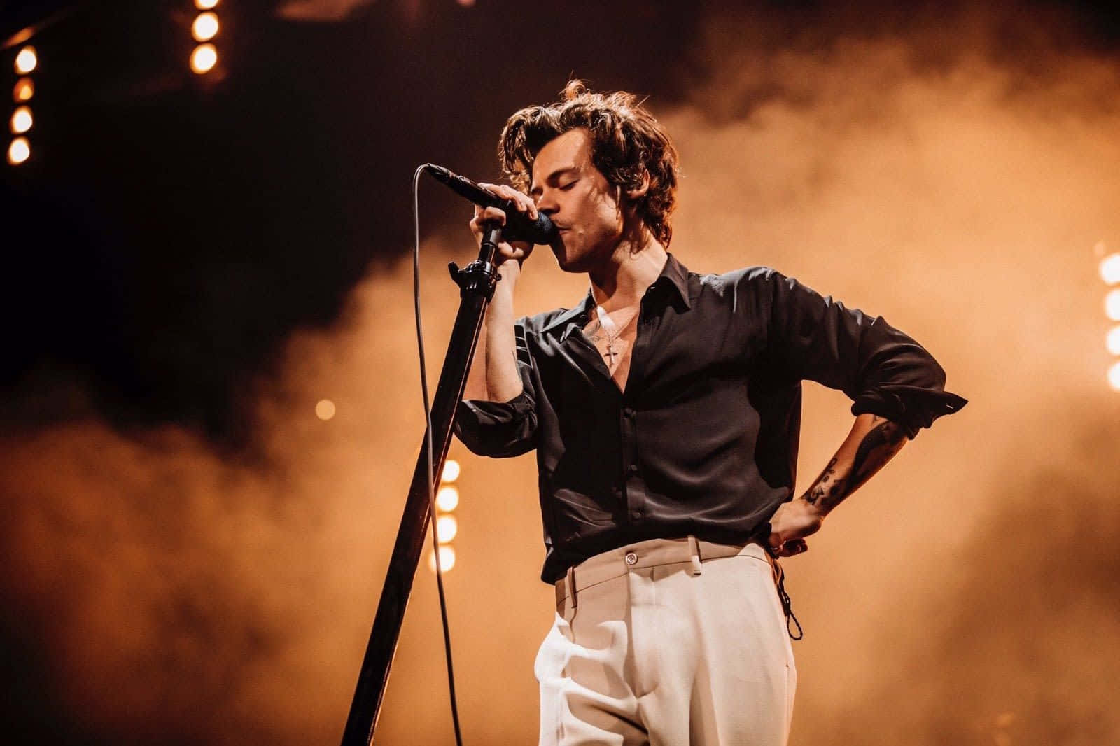 Harry Styles Laptop 桌布 壁紙 — 一位男歌手在溫暖的金色燈光和旋轉煙霧中熱情地在舞台上表演。