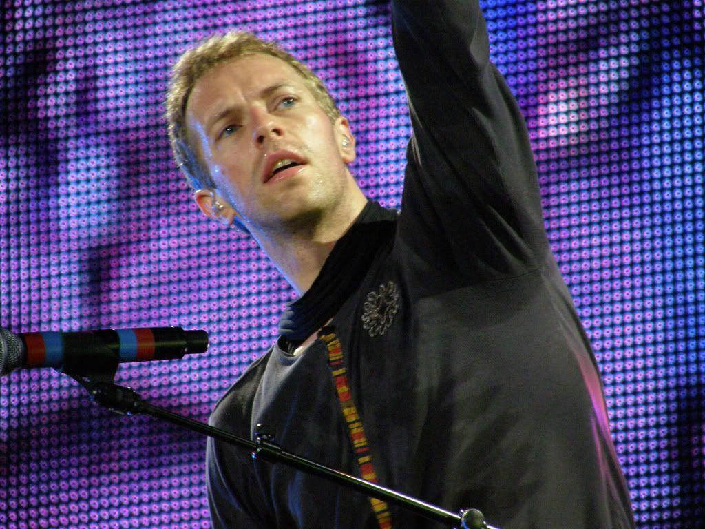 Chris Martin 桌布 壁紙 — 一位男歌手在舞台上熱情演出，單臂高舉，背景是充滿活力的紫色像素LED螢幕。