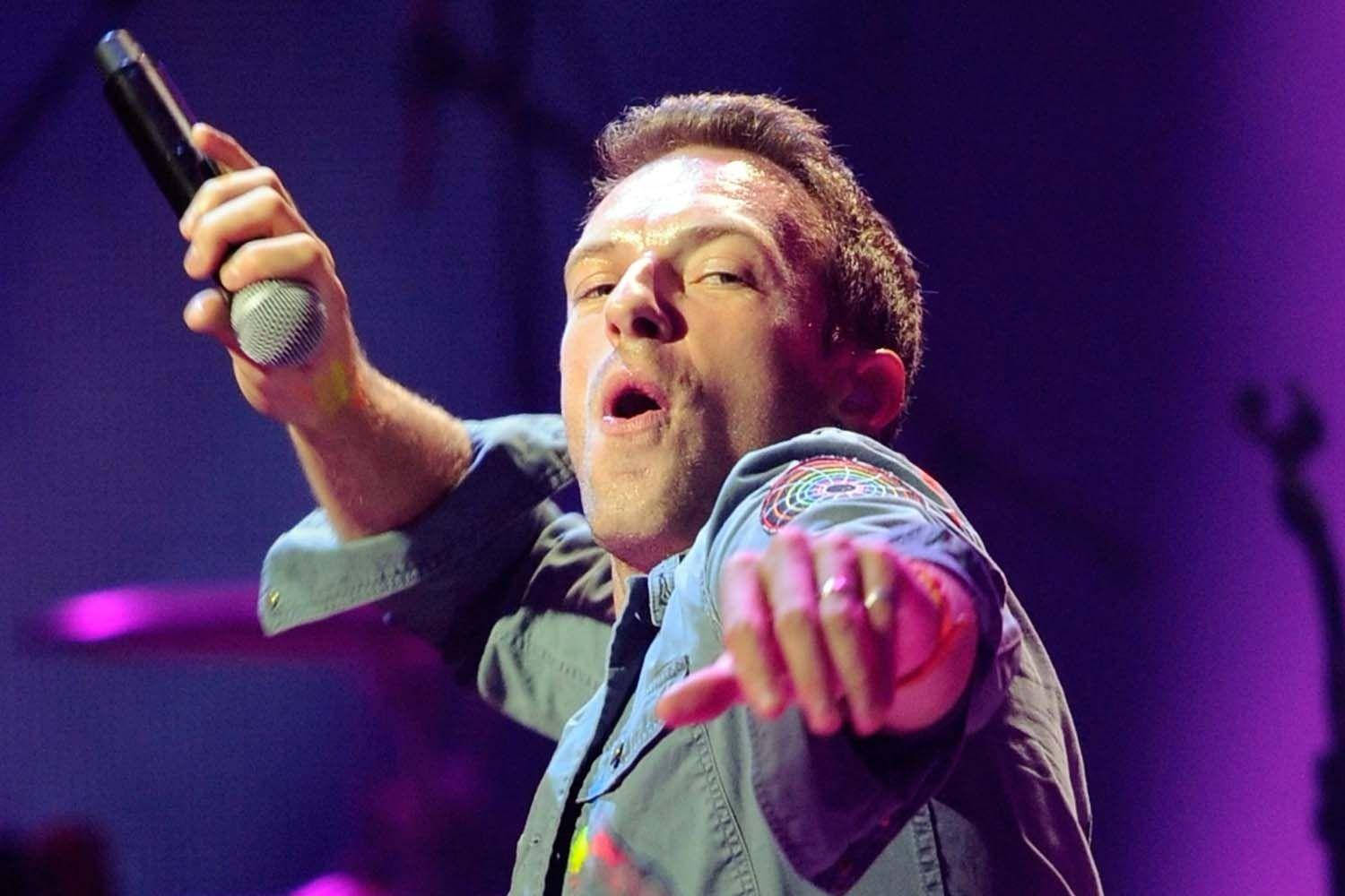 Chris Martin 桌布 壁紙 — 一位男歌手在舞台上熱情表演，手持麥克風，紫色舞台燈光照亮他的臉龐。