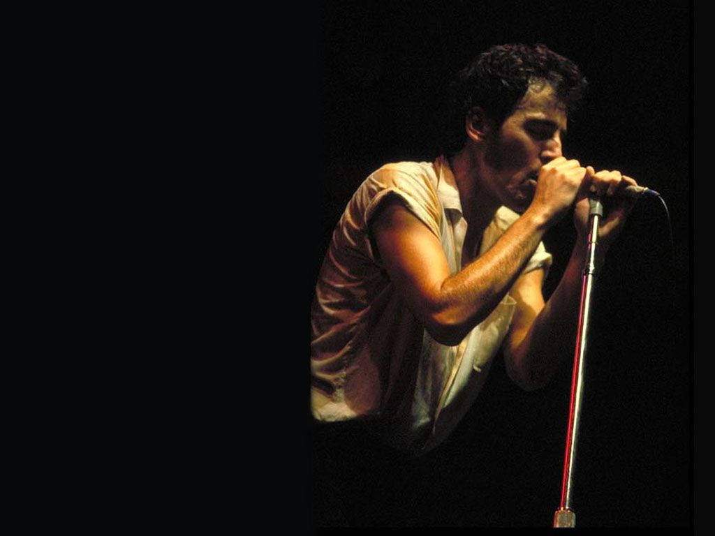 Bruce Springsteen 桌布 壁紙 — 一位男歌手在黑色背景前，於戲劇性的舞台燈光下熱情地對著麥克風演唱。