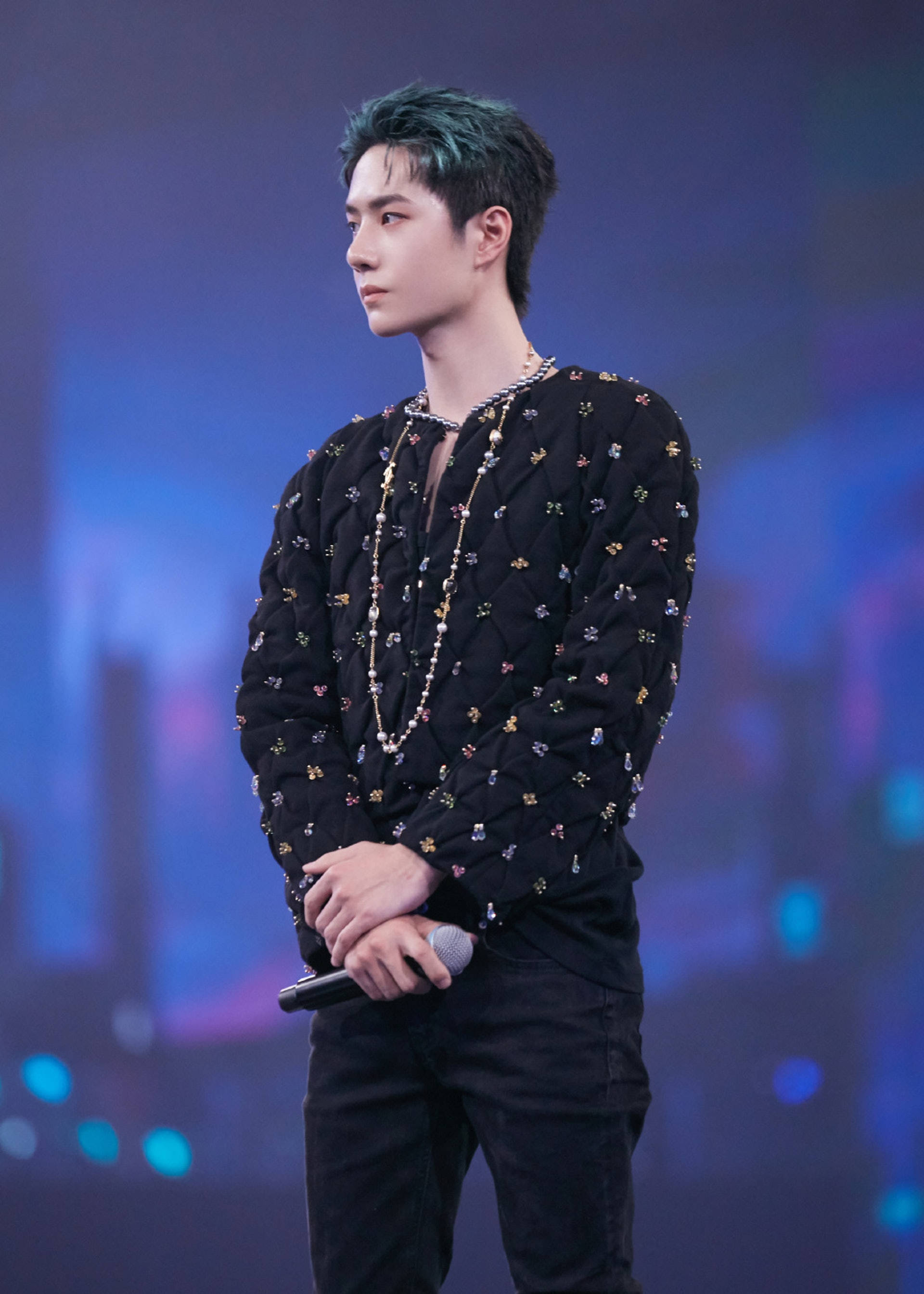 Wang Yibo 桌布 壁紙 — 一位男表演者帶有青綠色髮尖，身穿飾有彩色花卉刺繡的黑色絎縫外套，在藍色舞台燈光下拿著麥克風站在舞台上。