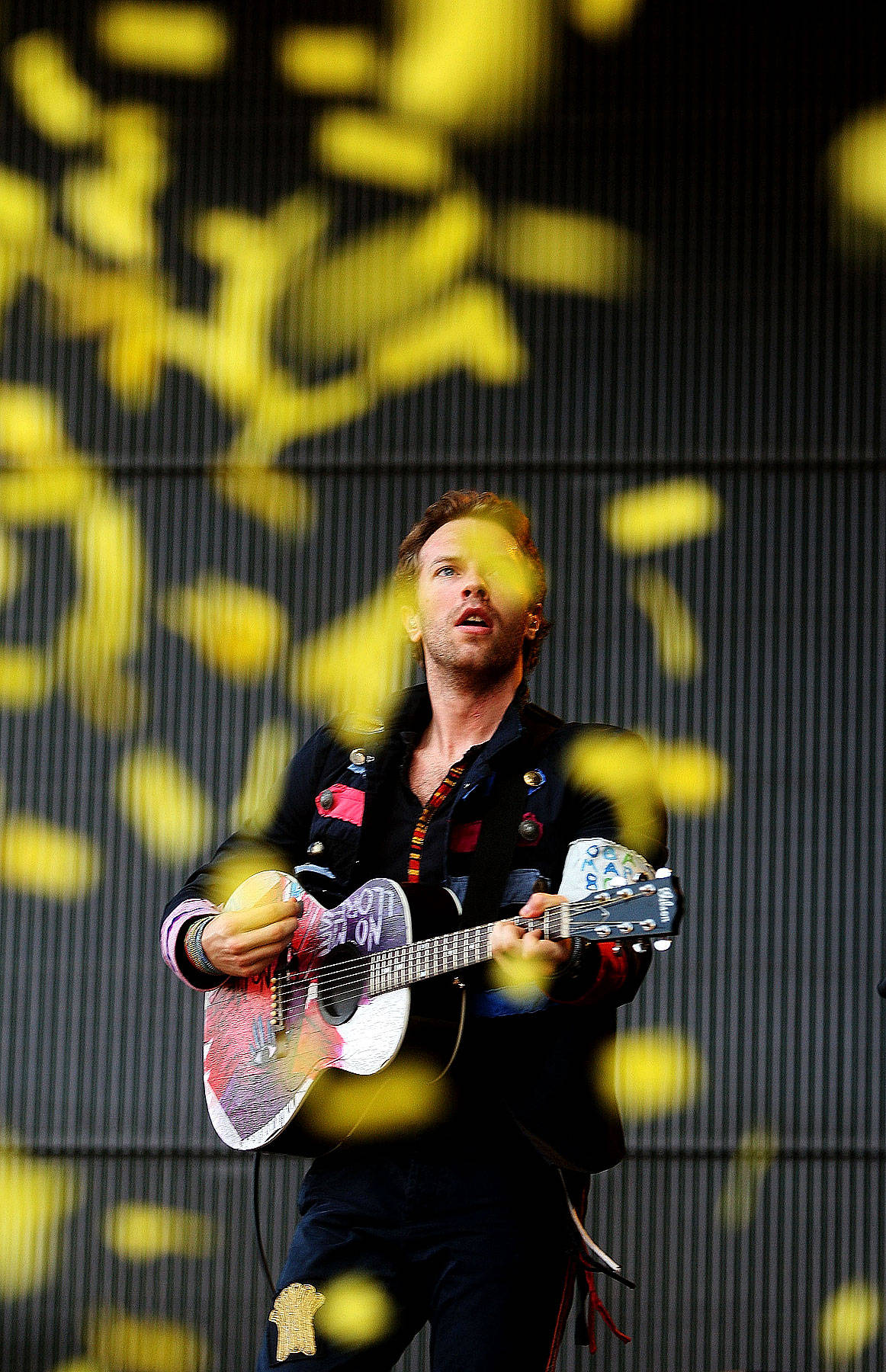 Chris Martin 桌布 壁紙 — 一位男音樂家在舞台上熱情地彈奏一把色彩繽紛的吉他，黃色紙屑如雨般落在他周圍。