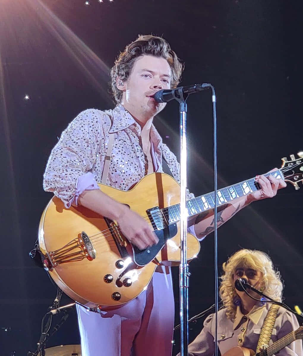 Harry Styles 2021 桌布 壁紙 — 一位男音樂家彈奏著一把旭日紋電吉他，在明亮的舞台燈光下對著麥克風唱歌，背景有一位樂隊成員。