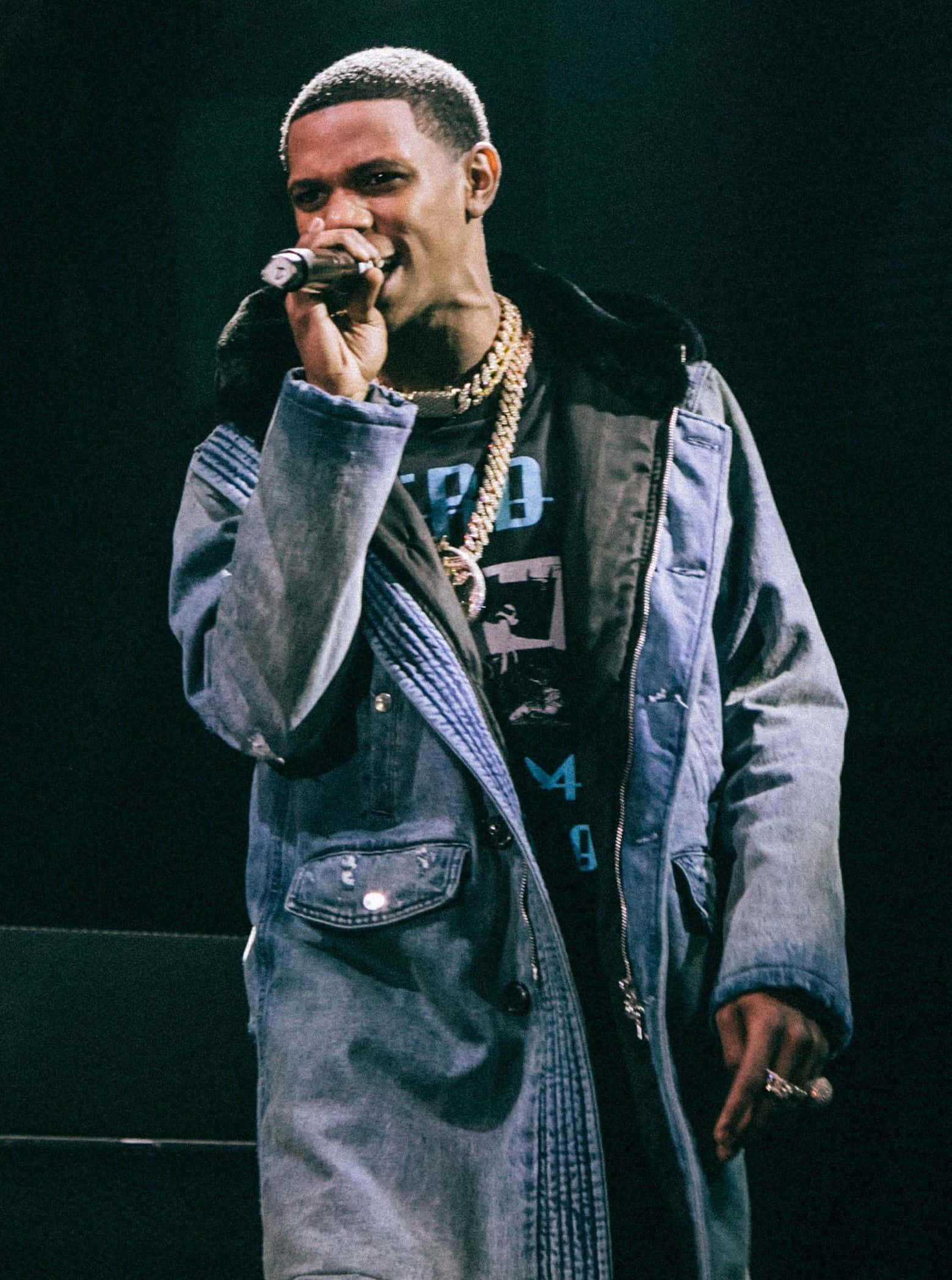 Rappers 桌布 壁紙 — 一位男饒舌歌手在舞台上表演，身穿牛仔外套和金色鏈條，在戲劇性的舞台燈光下拿著麥克風。
