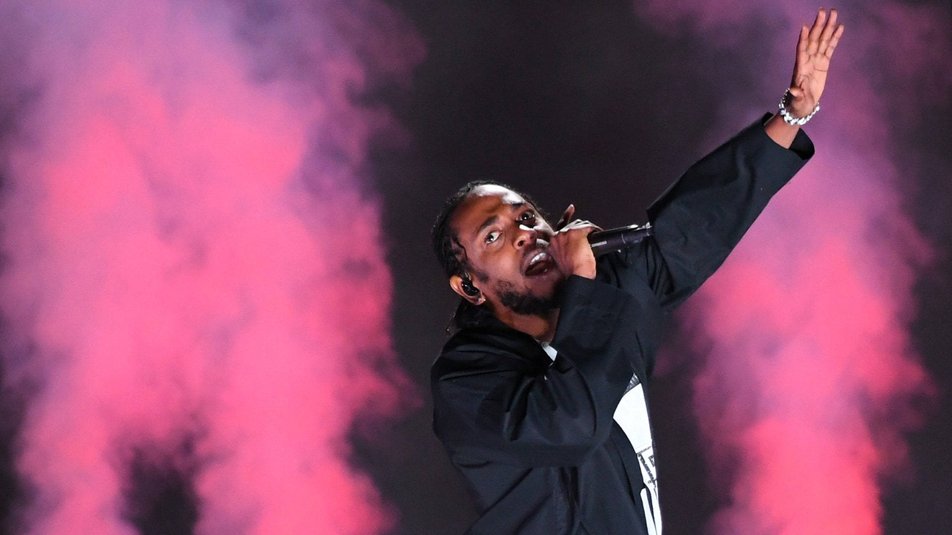 Kendrick 桌布 壁紙 — 一位男饒舌歌手在黑暗背景下，於充滿活力的粉紅色煙霧中熱情地舉起一隻手臂在舞台上表演。