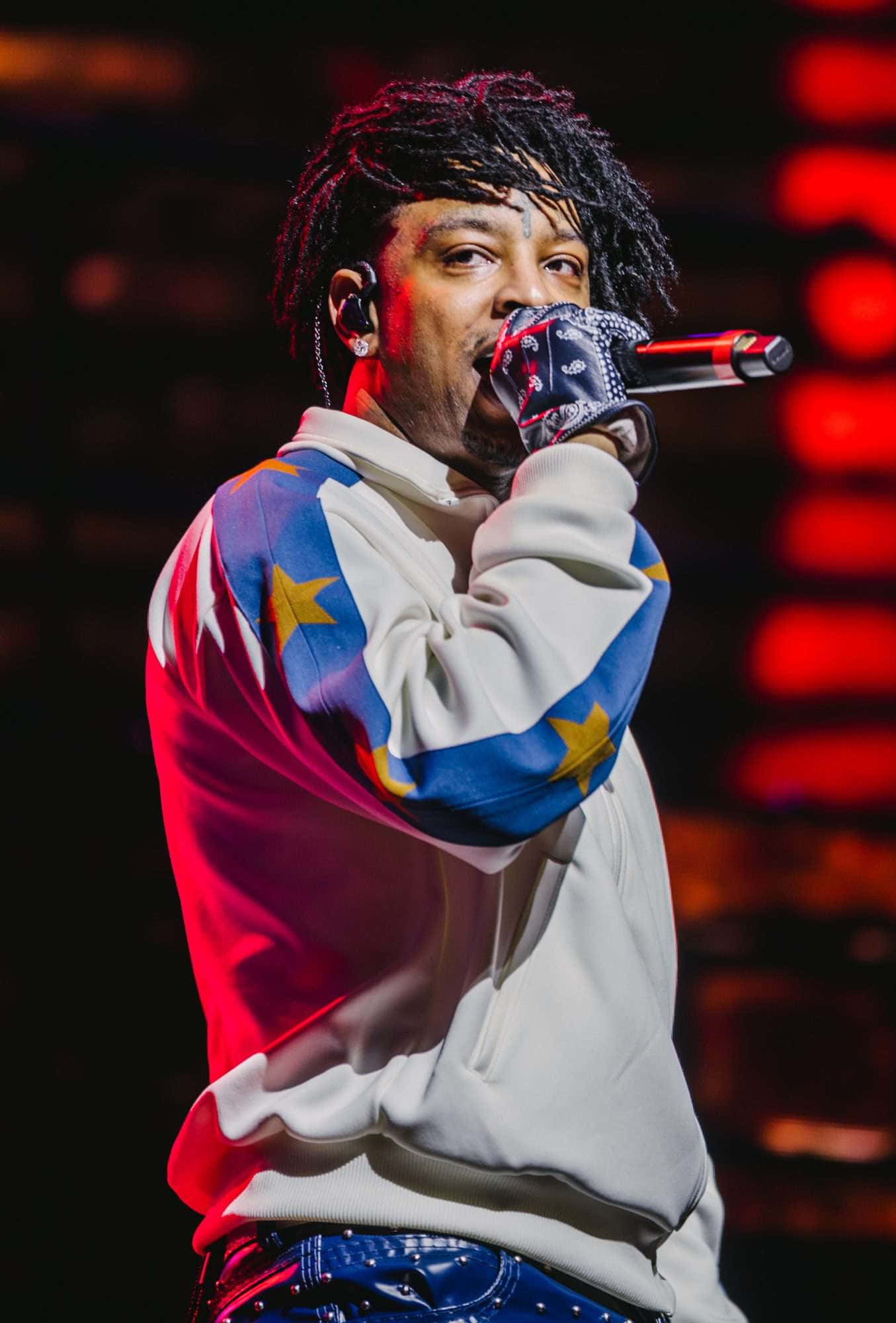 21 Savage 桌布 壁紙 — 一位男饒舌歌手梳著辮子，在紅色舞台燈光下，穿著星星圖案連帽衫，對著麥克風熱情表演。