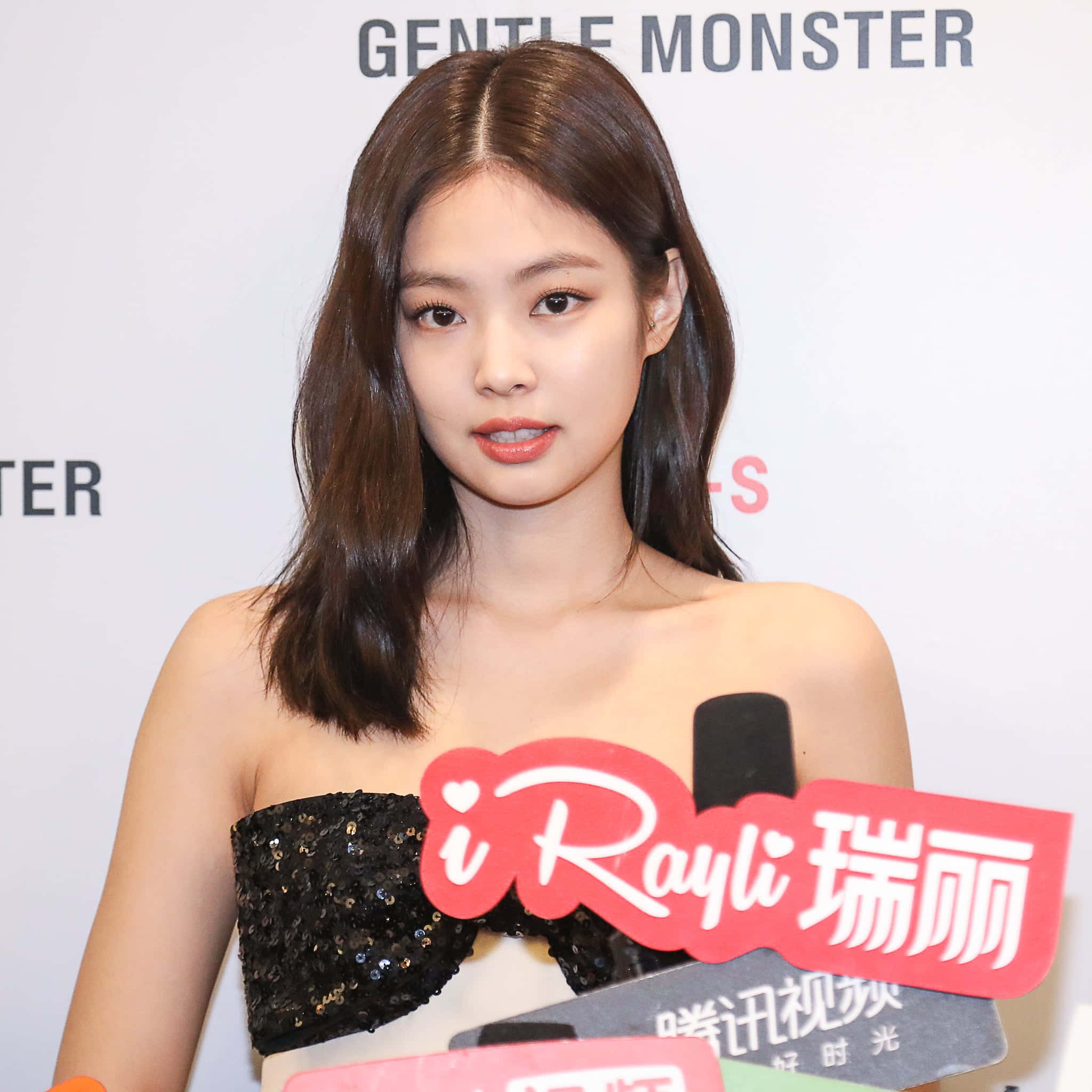 BLACKPINK 桌布 壁紙 — 一位留著及肩棕色長髮的女性，在設有麥克風和「GENTLE MONSTER」背景的記者會上，穿著黑色亮片無肩帶上衣。