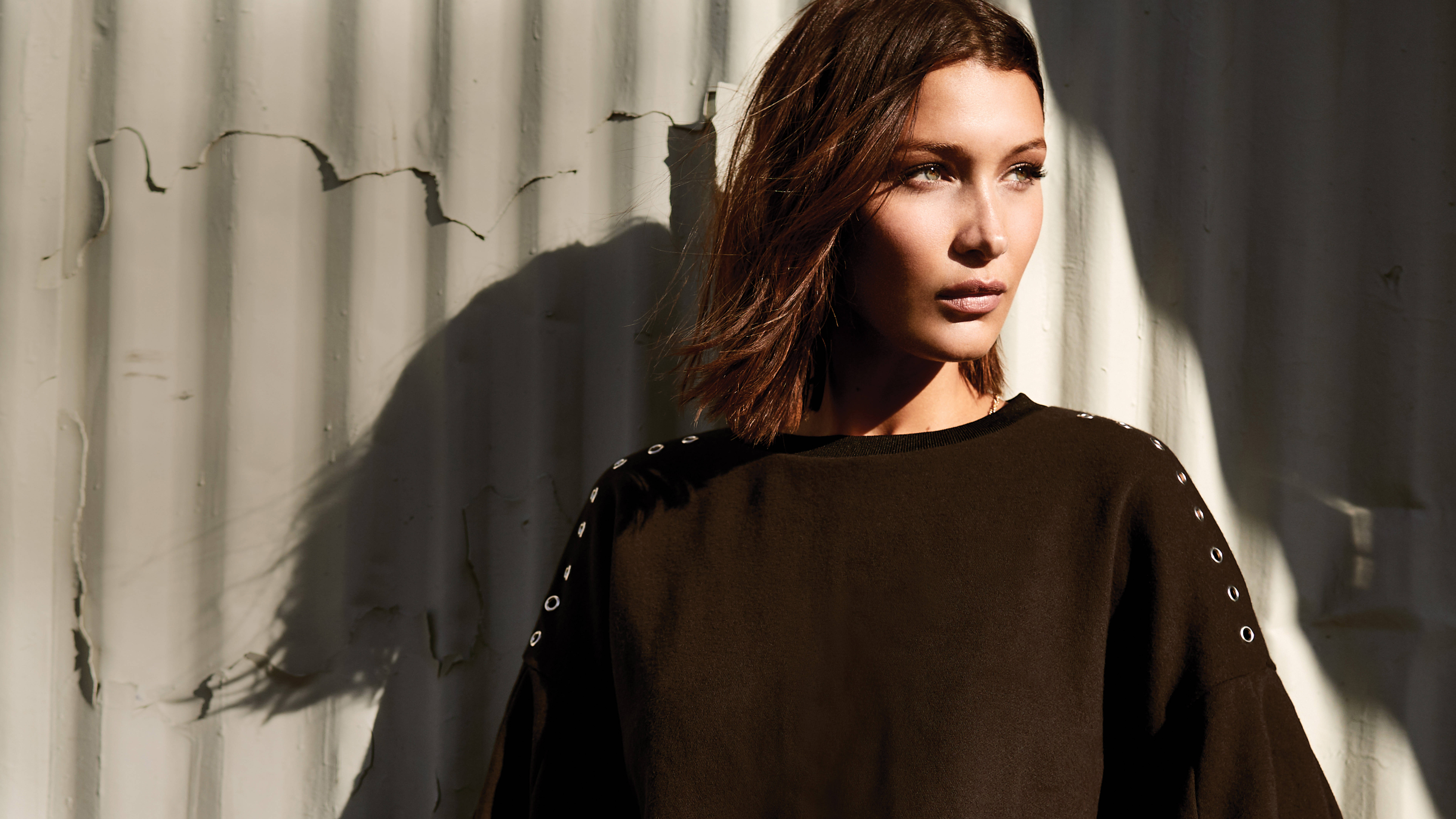 Bella Hadid 桌布 壁紙 — 一位留著及肩棕色頭髮的女性站在戲劇性的陽光下，陽光在她身後風化的波紋牆上投下銳利的陰影。