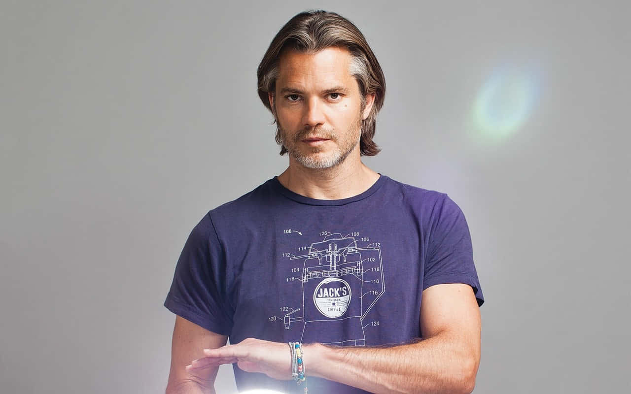 timothy olyphant 桌布 壁紙 — 一位留著及肩長髮和鬍鬚的男人，身穿印有咖啡機專利設計的紫色T恤，並用手比劃著。