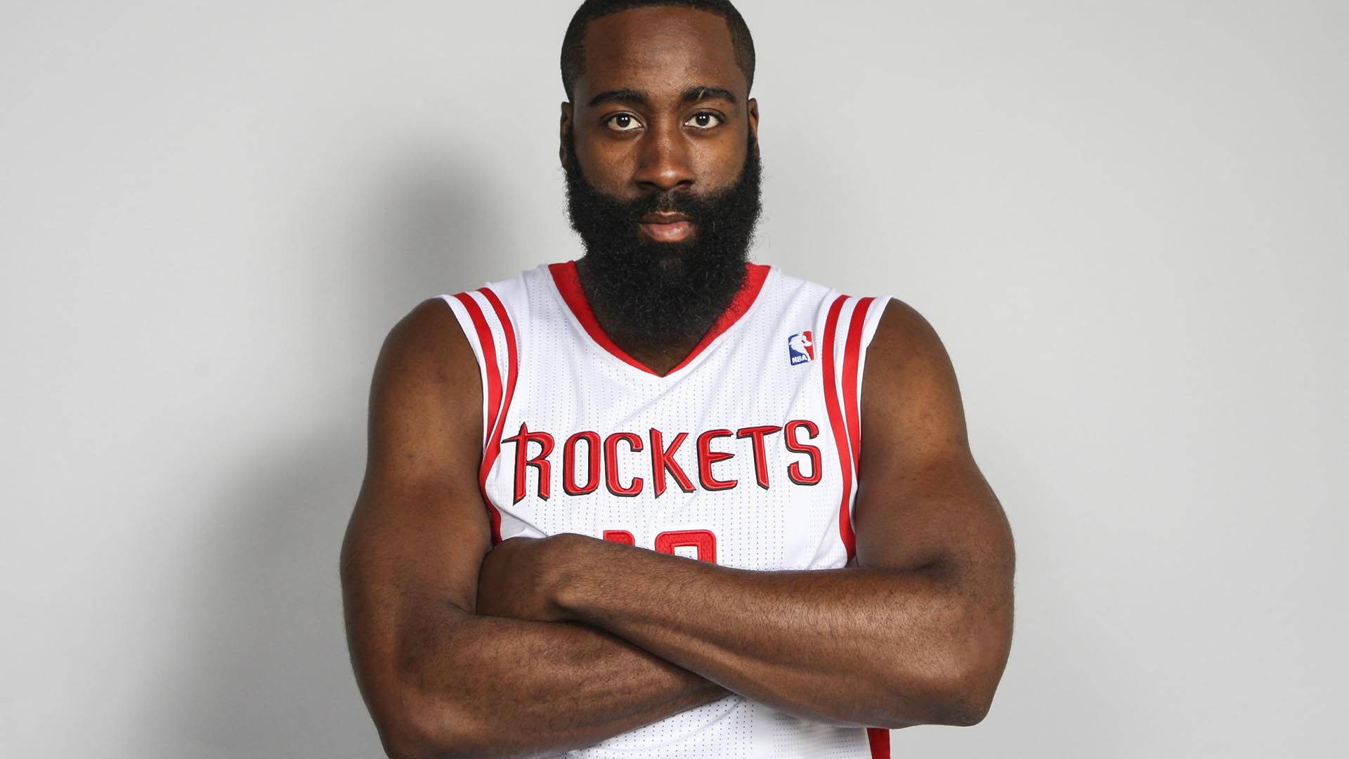 James Harden 桌布 壁紙 — 一位留著大鬍子的籃球運動員自信地穿著白色火箭隊球衣，雙臂交叉，站在純灰色背景前。
