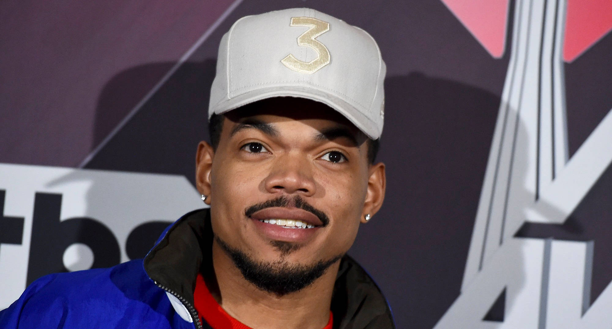 chance 桌布 壁紙 — 一位留著山羊鬍、戴著金色三號帽子的男士在品牌背景前對著鏡頭微笑。