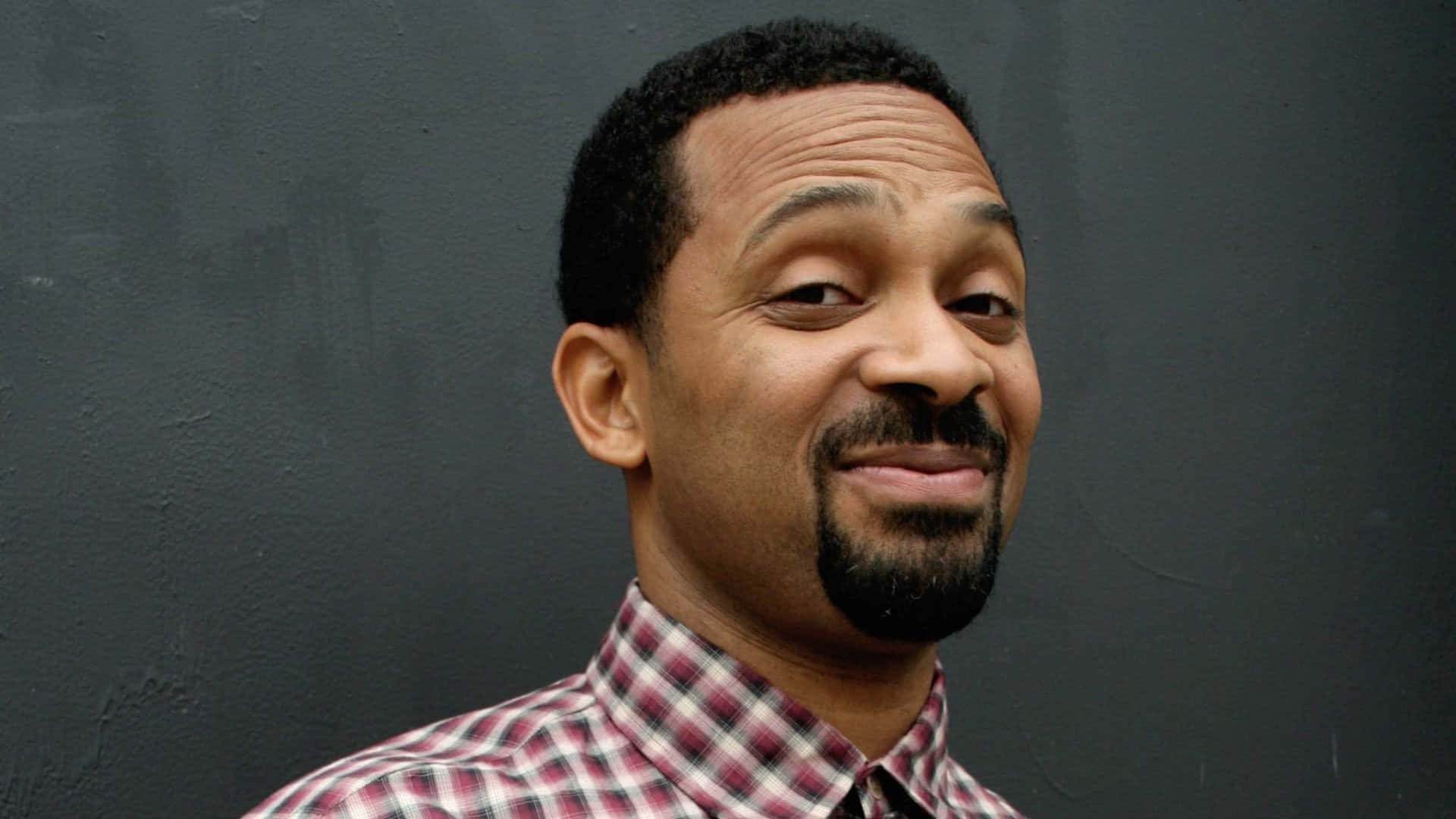 Mike Epps 桌布 壁紙 — 一位留著山羊鬍、短髮的男子，穿著紅白格紋襯衫，在深灰色紋理牆前溫暖微笑。
