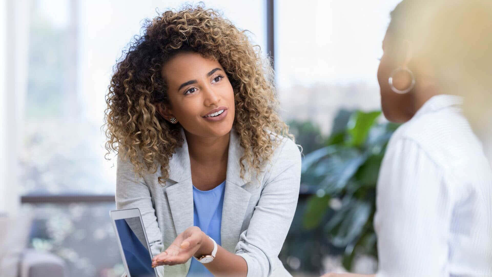 professional 桌布 壁紙 — 一位留著捲髮、身穿灰色西裝外套的女性，在明亮的辦公室環境中與同事交談時做手勢。