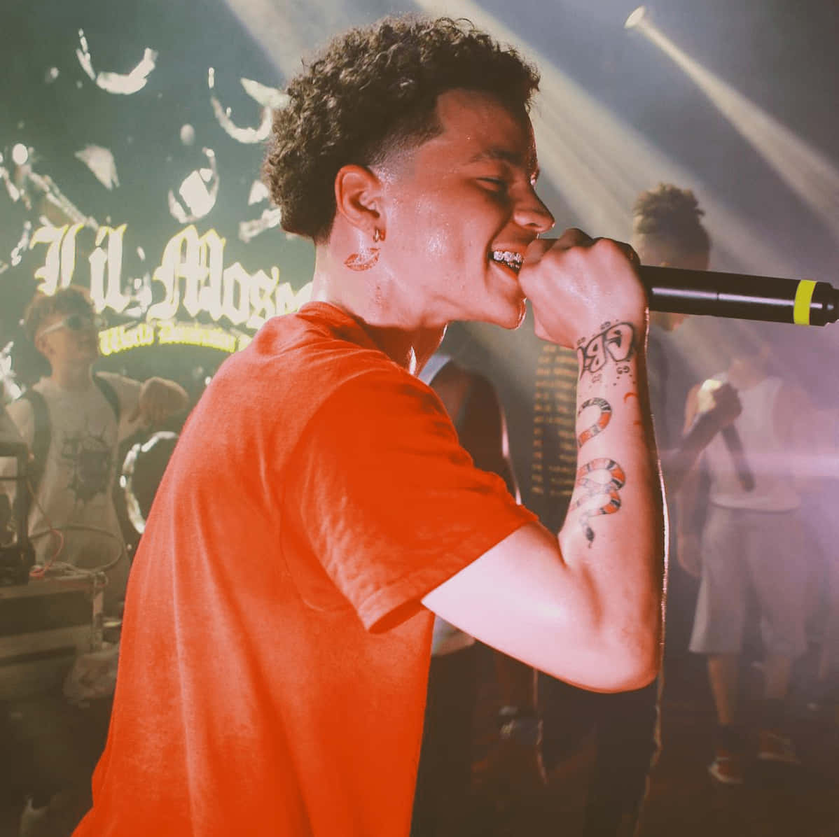 Lil Mosey 桌布 壁紙 — 一位留著捲髮和刺青的年輕男子在現場演唱會的舞台燈光下，熱情地對著麥克風唱歌。