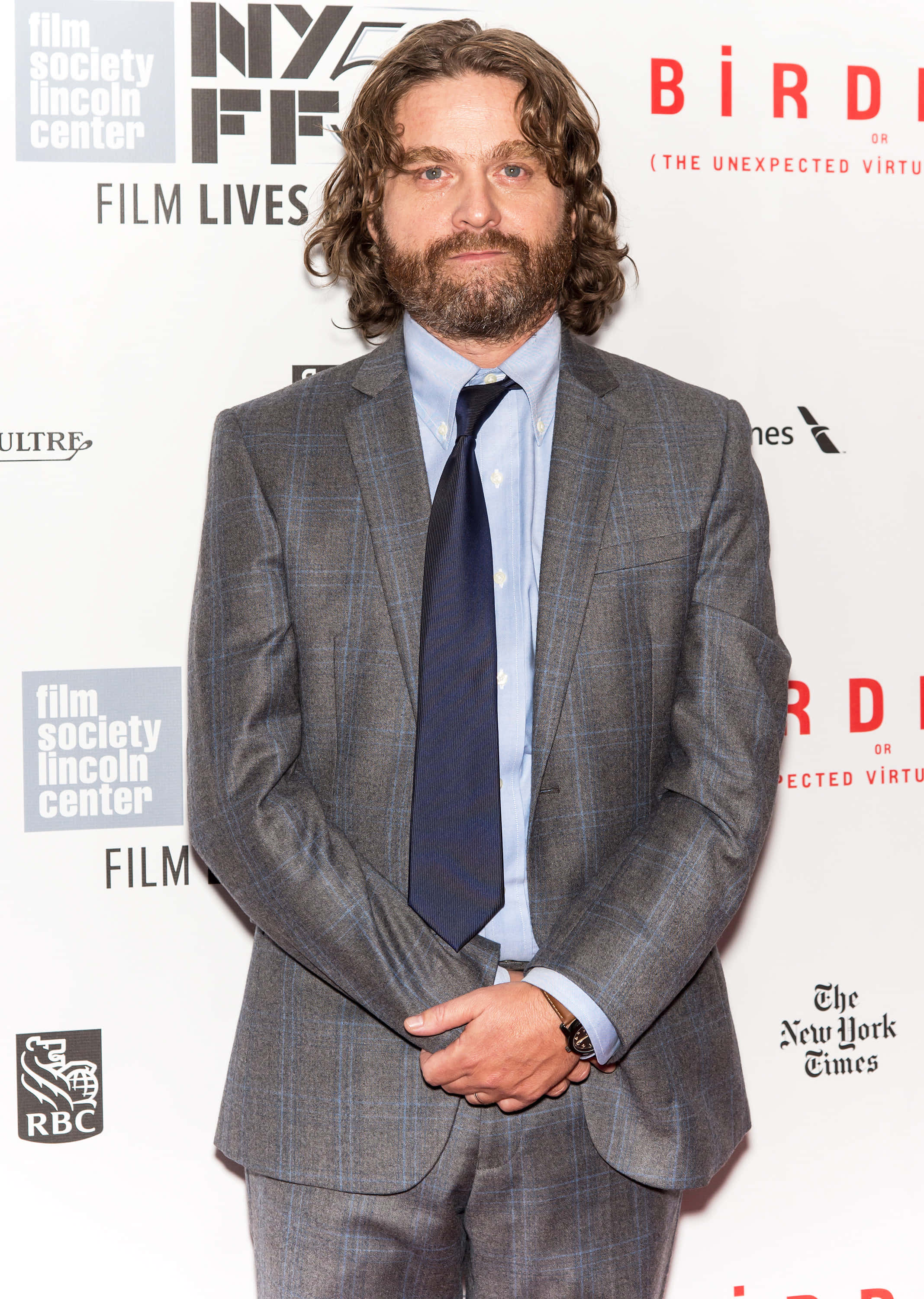 Zach Galifianakis 桌布 壁紙 — 一位留著捲髮和鬍鬚的男子，身穿灰色格紋西裝，自信地站在電影節活動現場。