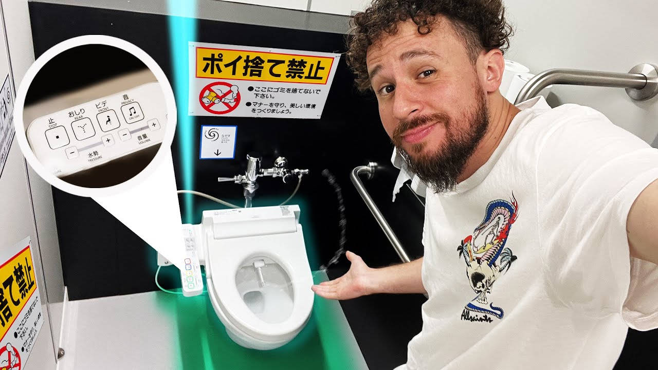 日本 桌布 壁紙 — 一位留著捲髮和鬍鬚的男子微笑著指向一個帶有綠色發光裝飾和操控面板的高科技日本馬桶。