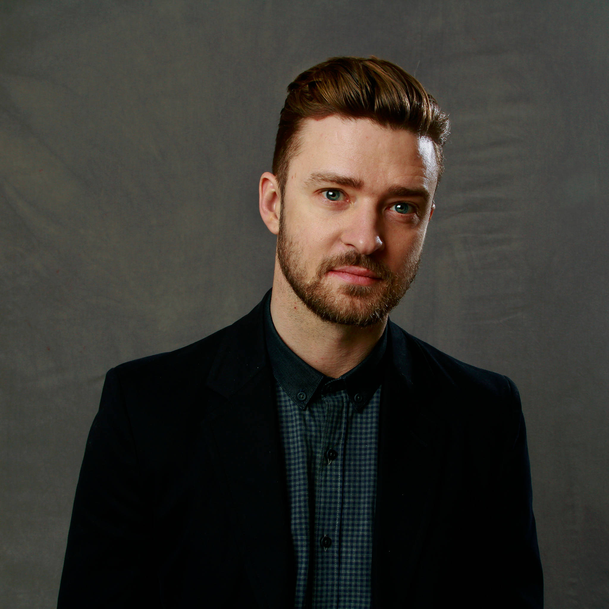 justin timberlake 桌布 壁紙 — 一位留著時尚棕色頭髮和鬍鬚的男子，身穿深色西裝外套，內搭格紋襯衫，背景為有紋理的灰色。