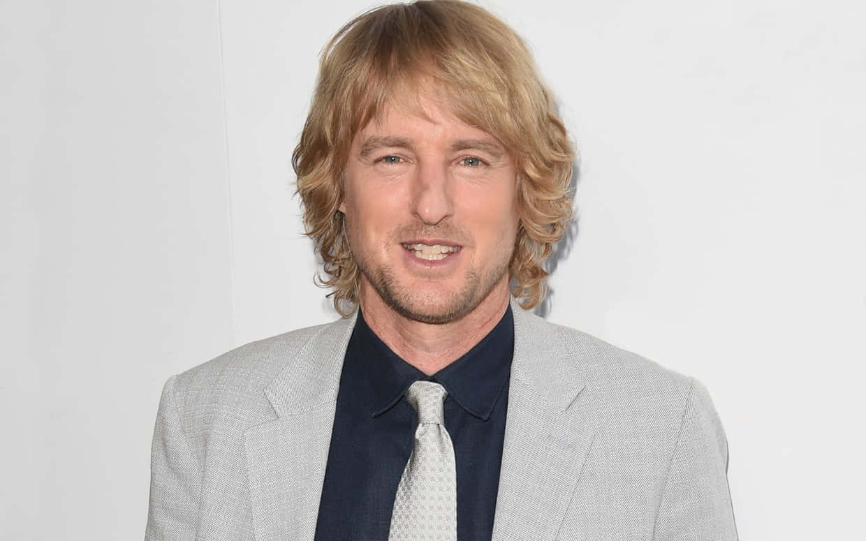 Owen Wilson 桌布 壁紙 — 一位留著波浪形金髮、身穿淺灰色西裝的男子在純白背景前微笑。