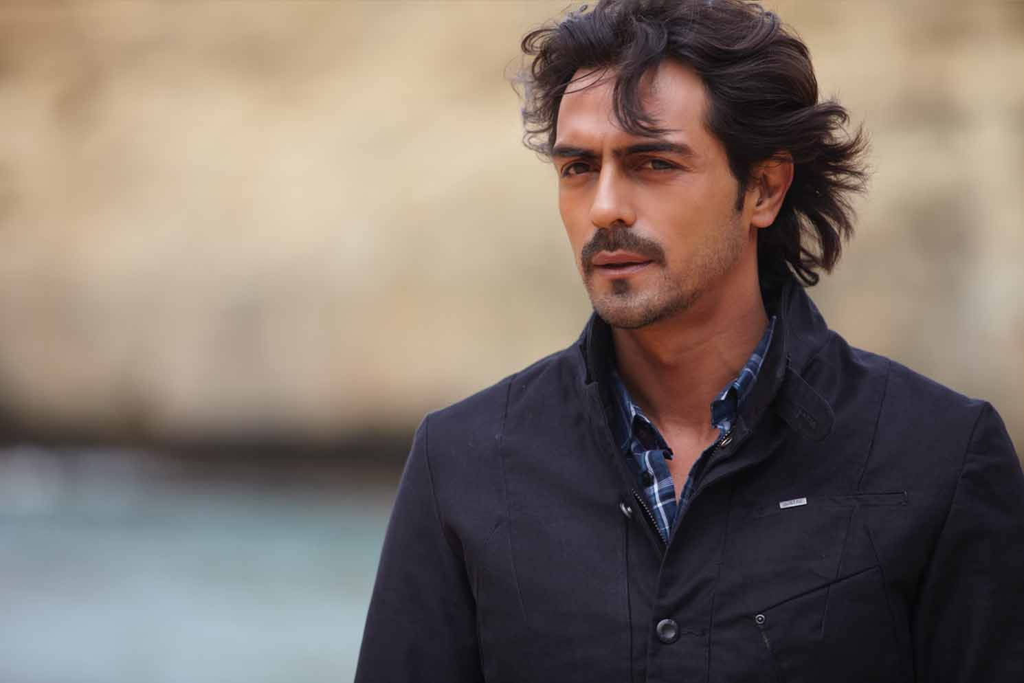 Arjun Rampal 桌布 壁紙 — 一位留著深色波浪髮和鬍鬚的男子，身穿深色外套內搭格紋襯衫，站在水邊。