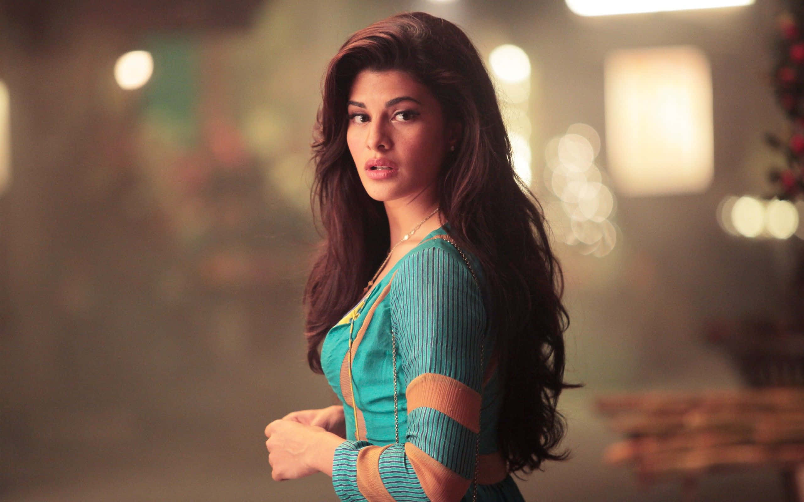 Jacqueline Fernandez 桌布 壁紙 — 一位留著深色長髮、身穿青綠色羅紋連衣裙的女性，在溫暖照明的室內環境中轉頭越過肩膀回望。