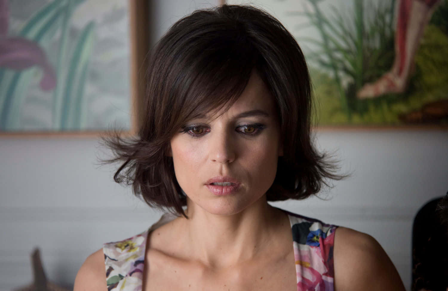 Elena Anaya 桌布 壁紙 — 一位留著深色鮑伯頭、身穿花卉圖案上衣的女性，在柔和光線的室內環境中向下凝視。