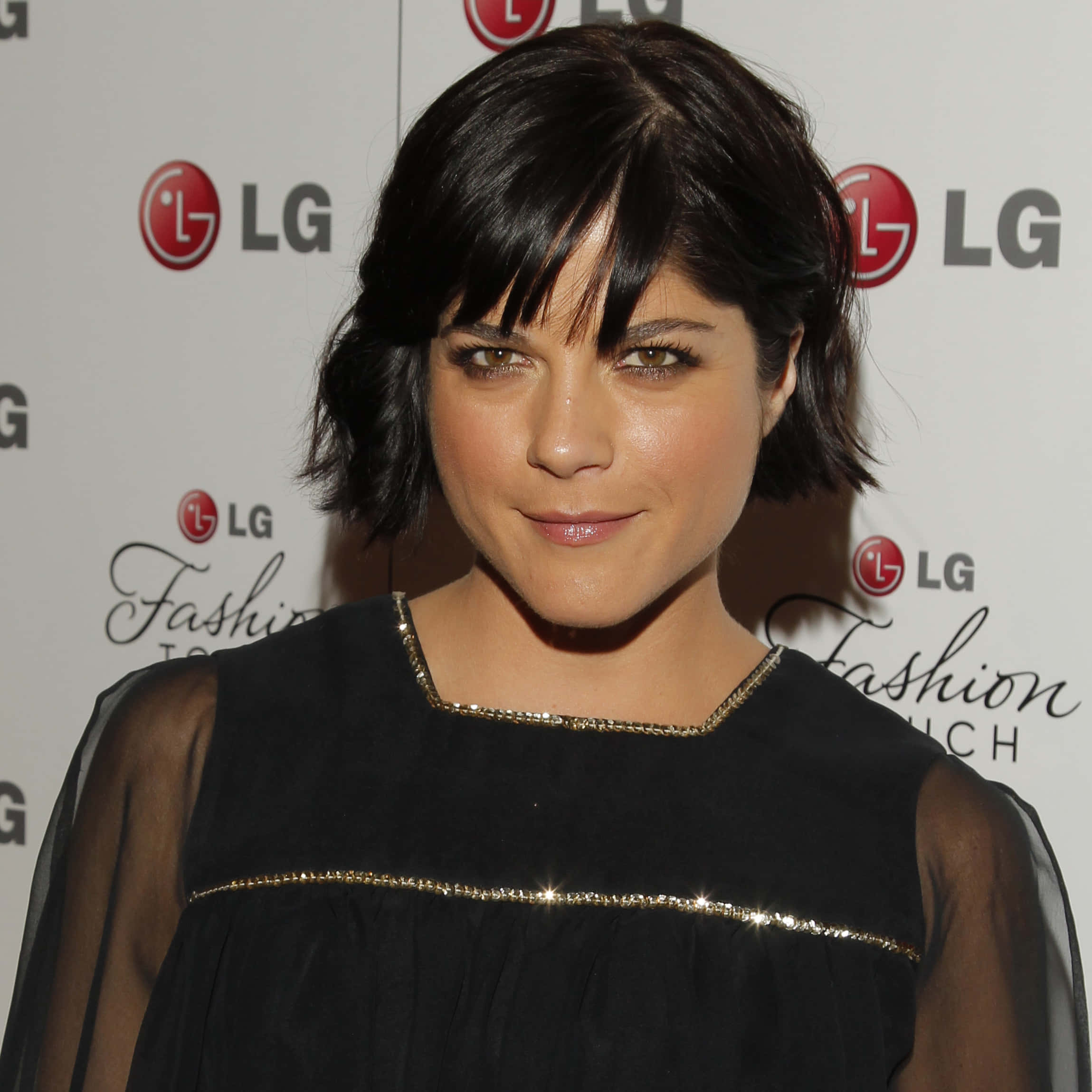 Selma Blair 桌布 壁紙 — 一位留著深色鮑伯頭和瀏海的女性，在LG Fashion Touch活動中穿著帶有金色飾邊的黑色連衣裙，溫柔地微笑。