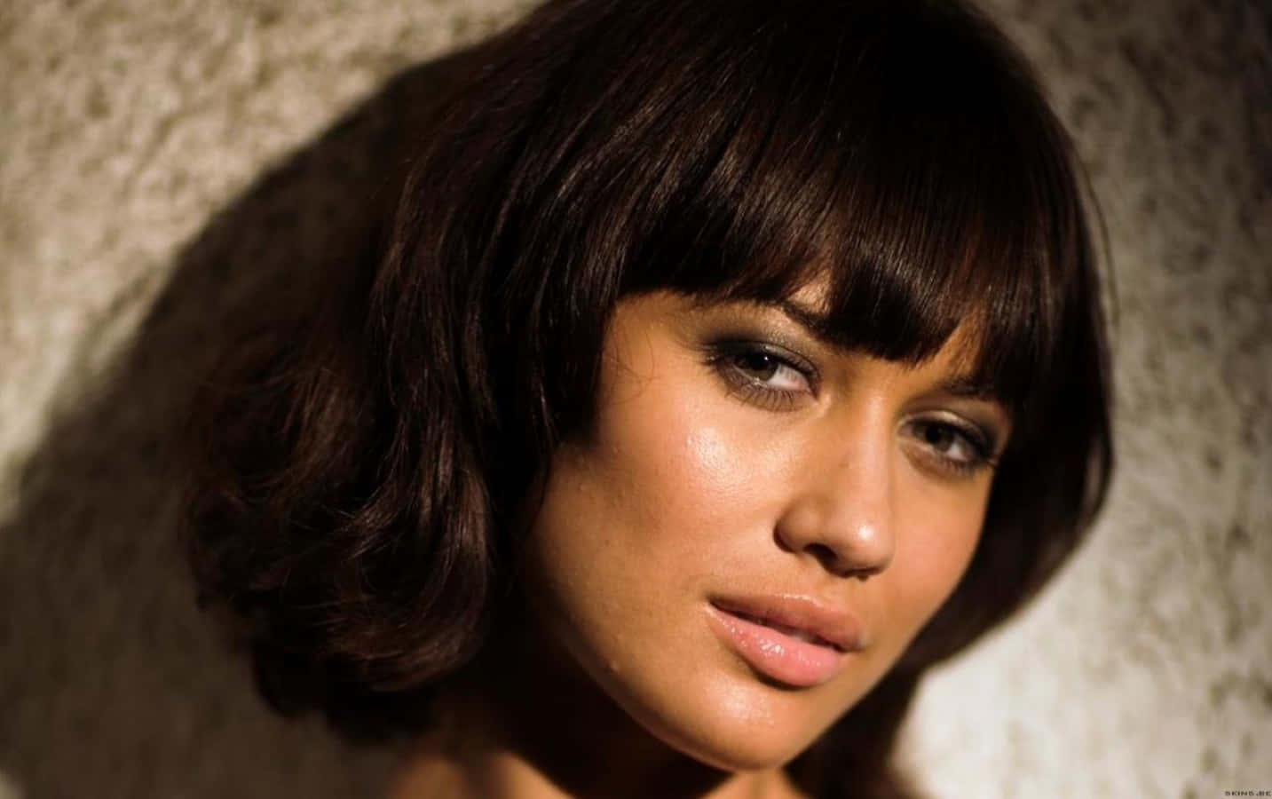 Olga Kurylenko 桌布 壁紙 — 一位留著深色鮑伯頭和瀏海的女性，在溫暖的方向性燈光下，柔和地凝視著一面有紋理的牆壁。