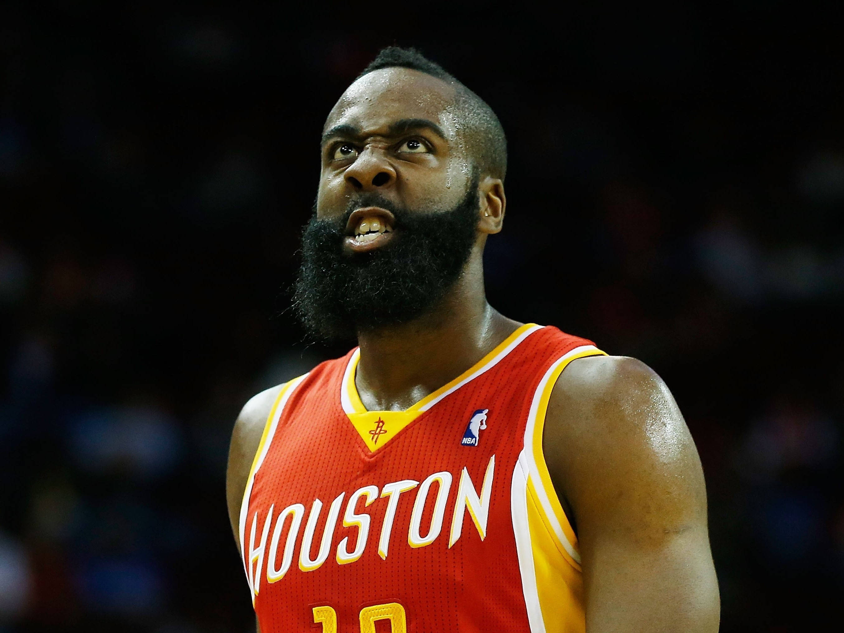 Houston Rockets 桌布 壁紙 — 一位留著濃密鬍鬚、專注的籃球運動員，在明亮的場館燈光下，於一場激烈的比賽中穿著紅色休士頓火箭隊球衣。