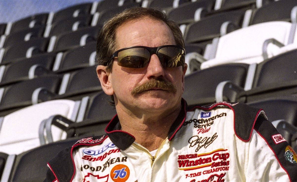 Sunglasses 桌布 壁紙 — 一位留著濃密鬍鬚、戴著太陽眼鏡的NASCAR車手，在明亮的日光下，穿著賽車服，以空蕩蕩的體育場座位為背景擺姿勢。