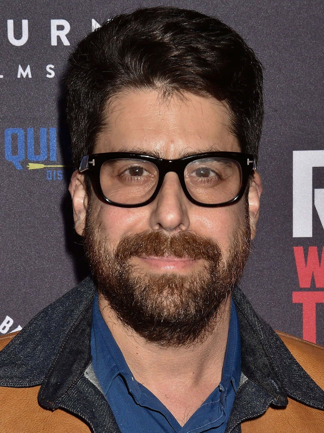actor 桌布 壁紙 — 一位留著濃密鬍鬚、戴著粗黑框眼鏡的男人對著鏡頭微笑，身穿牛仔外套內搭藍色襯衫。