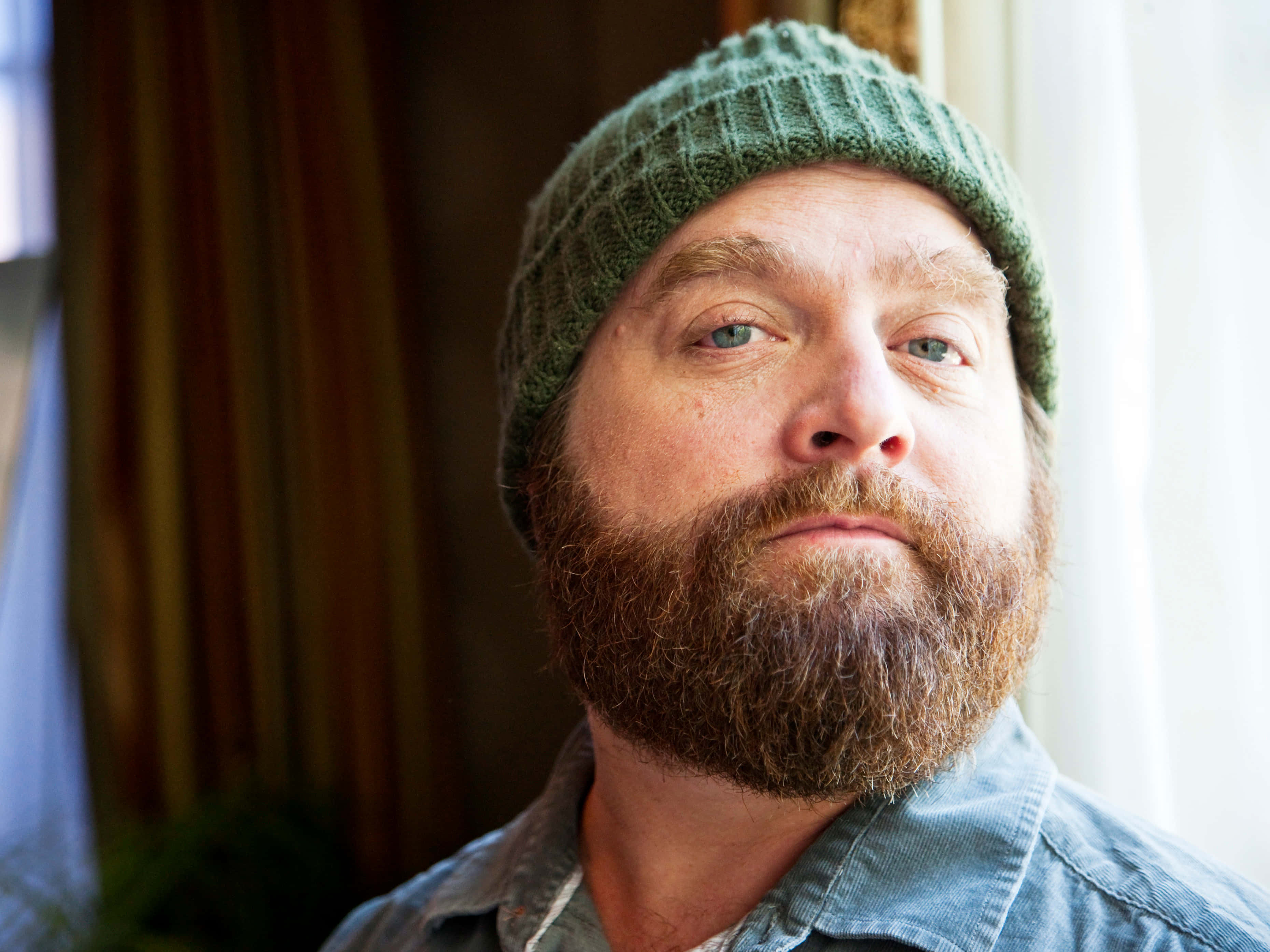 Zach Galifianakis 桌布 壁紙 — 一位留著濃密鬍鬚、戴著綠色針織毛帽的男子，在柔和自然光下若有所思地凝視鏡頭。