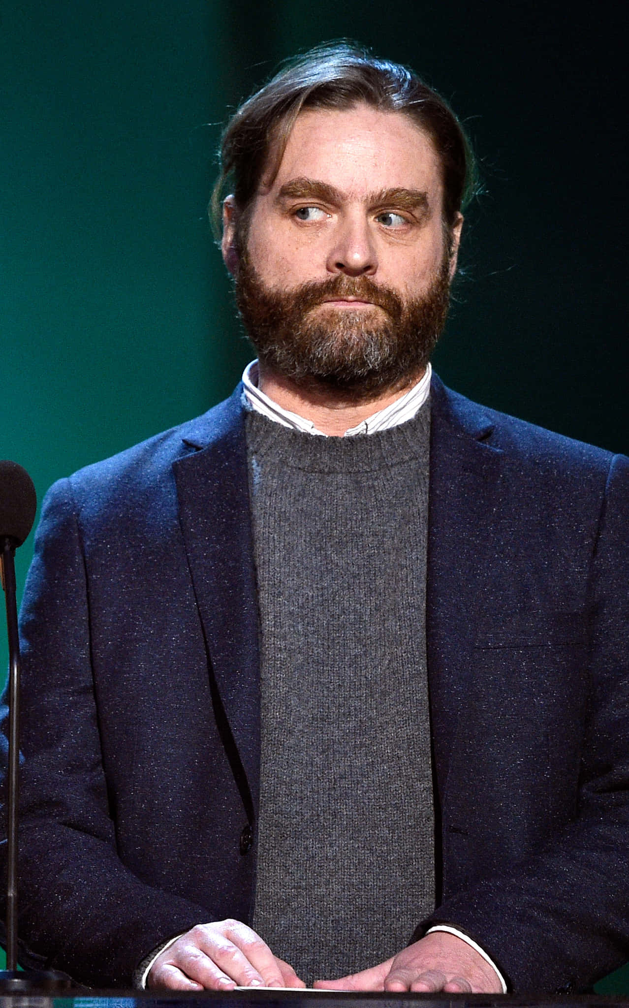 Zach Galifianakis 桌布 壁紙 — 一位留著濃密鬍鬚、深色頭髮的男子站在講台上，身穿海軍藍西裝外套，內搭灰色毛衣，背景為綠色。