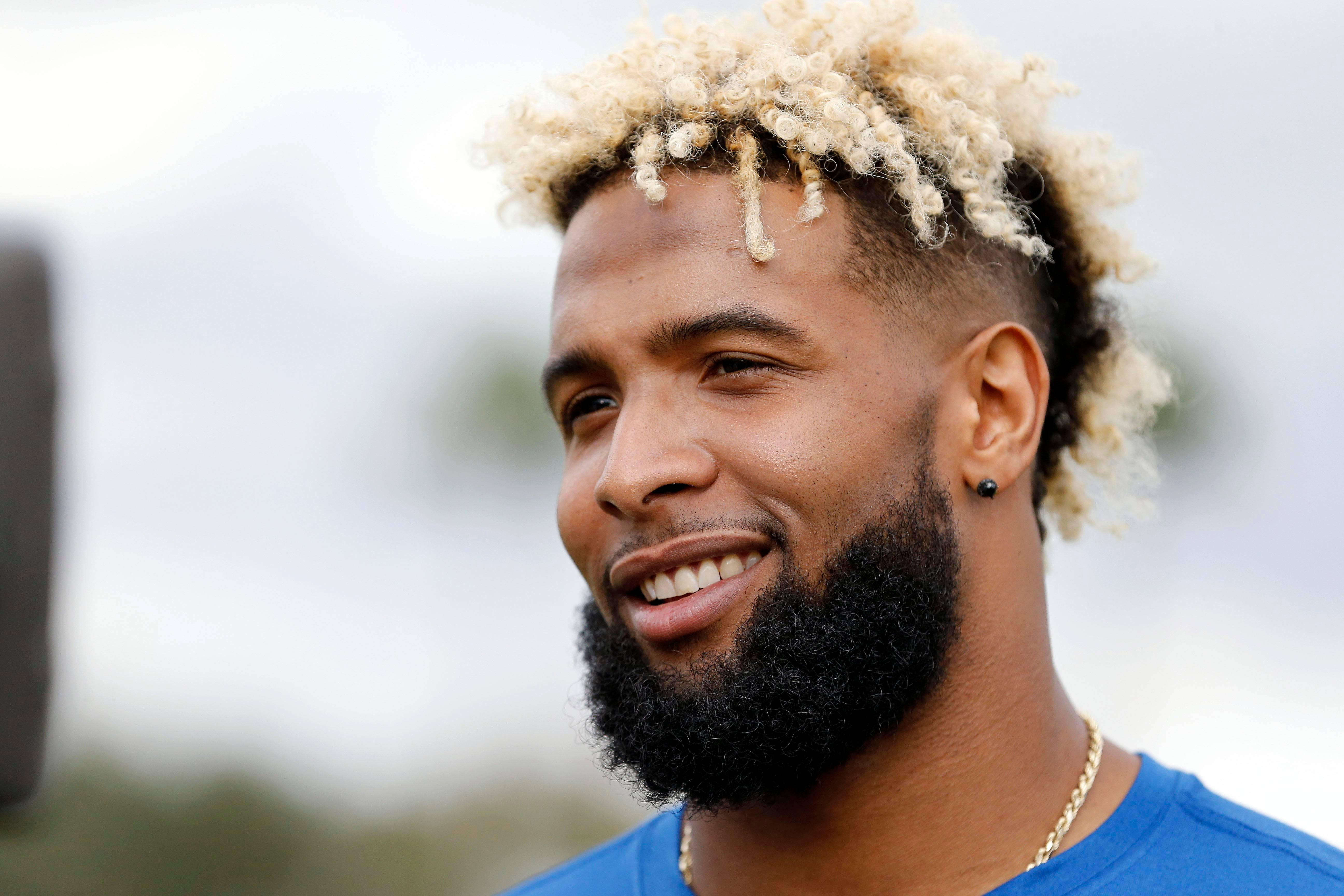 Odell 桌布 壁紙 — 一位留著濃密鬍鬚、漂白捲髮的微笑男子，身穿藍色襯衫和金色項鍊，背景是柔和模糊的戶外景色。