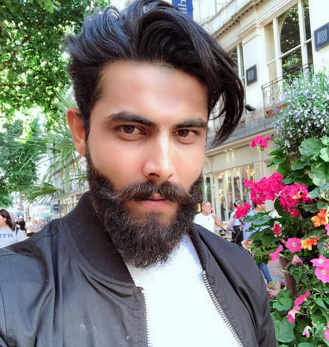 Indian 桌布 壁紙 — 一位留著濃密鬍鬚和時尚髮型的男士，在陽光明媚的城市街道上自拍，街道兩旁盛開著鮮豔的粉紅色花朵和古典建築。