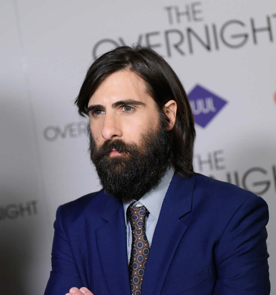 Jason Schwartzman 桌布 壁紙 — 一位留著濃密鬍鬚和長黑髮的男子，在推廣活動中穿著俐落的藍色西裝和圖案領帶。