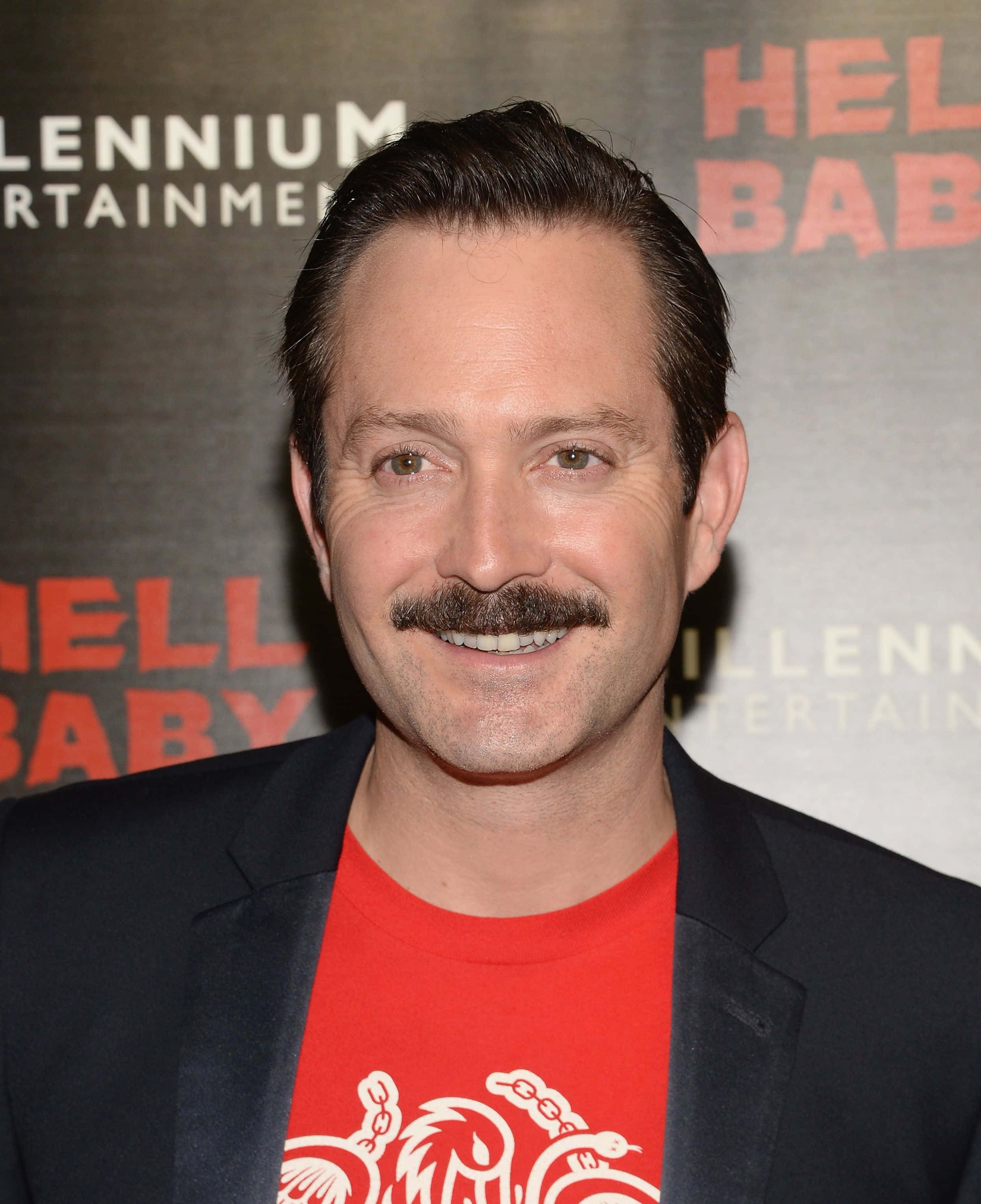 thomas lennon 桌布 壁紙 — 一位留著濃密鬍鬚的微笑男士，在電影首映活動中穿著黑色西裝外套內搭紅色T恤。