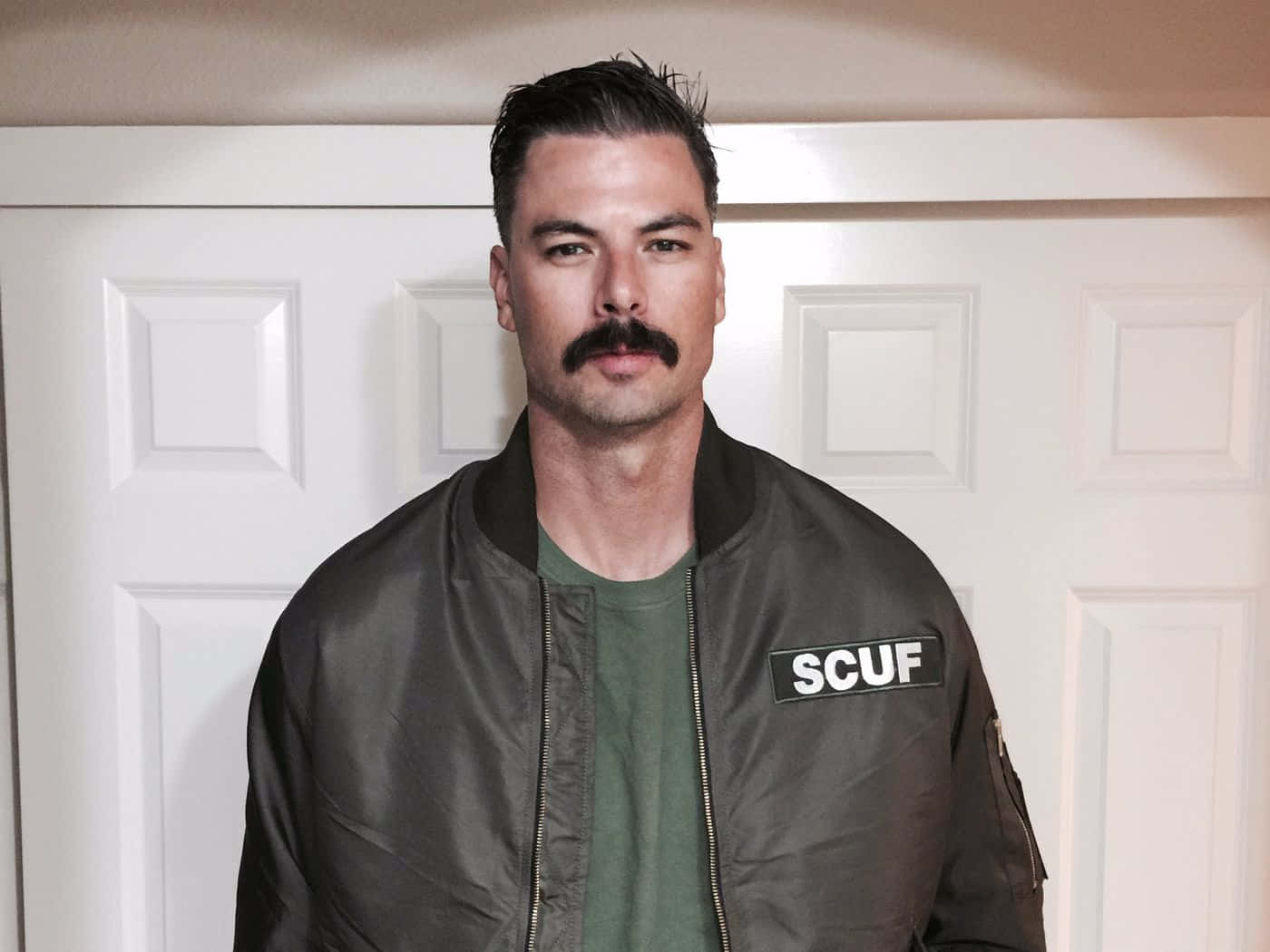 Dr Disrespect 桌布 壁紙 — 一位留著濃密鬍鬚的男子，身穿帶有SCUF補丁的深色轟炸機夾克，站在白色鑲板門前。
