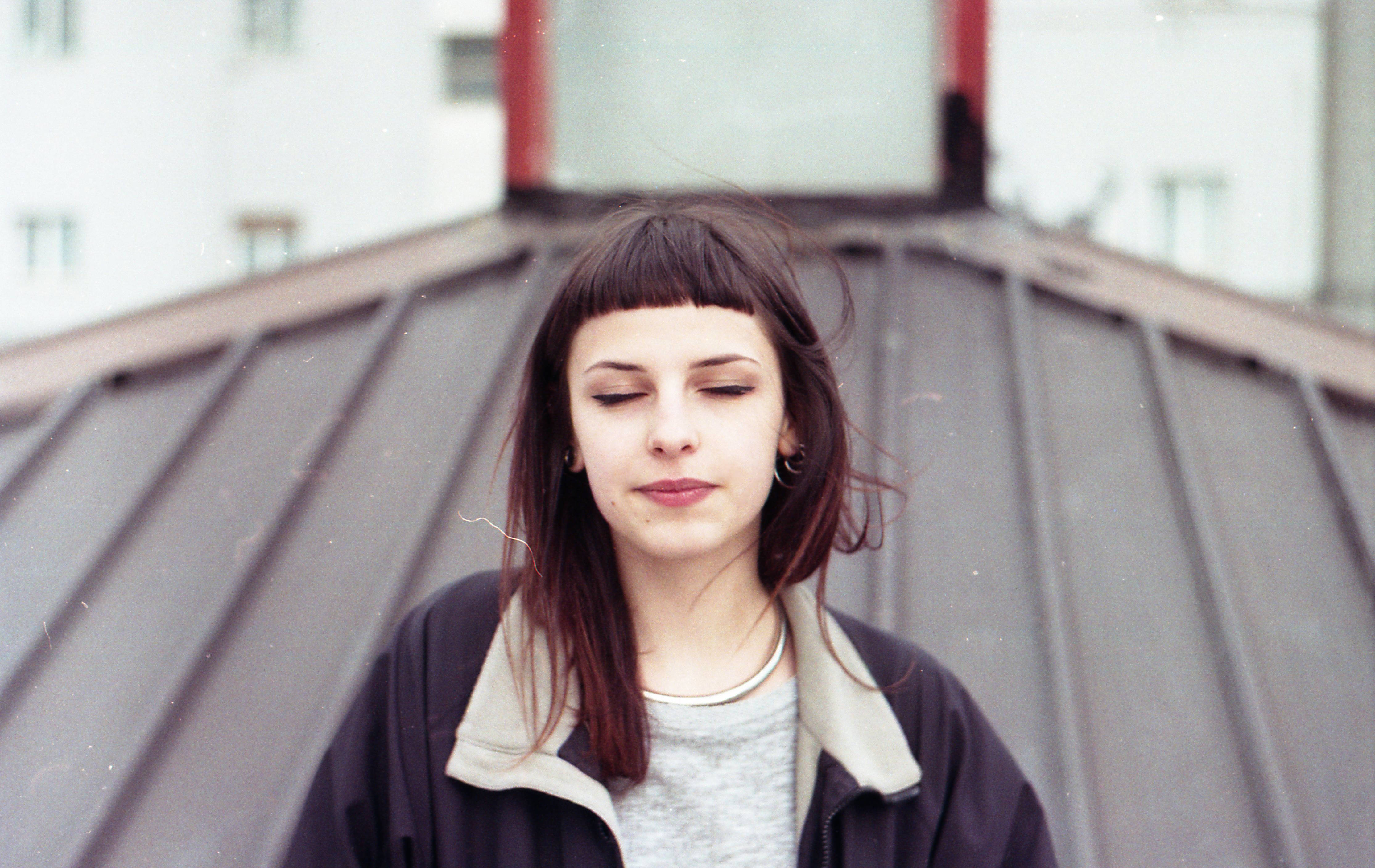 美女 桌布 壁紙 — 一位留著瀏海的年輕女子平靜地站在金屬屋頂上，身後建築物模糊不清。