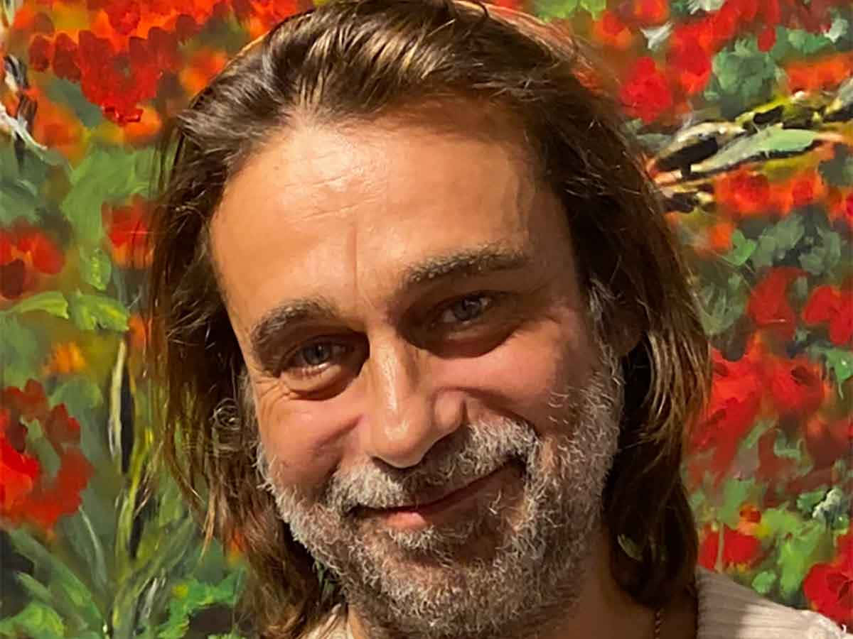 Jordi Mollà 桌布 壁紙 — 一位留著灰白鬍子的男士在鮮豔的紅綠色花卉畫作前溫暖地微笑。