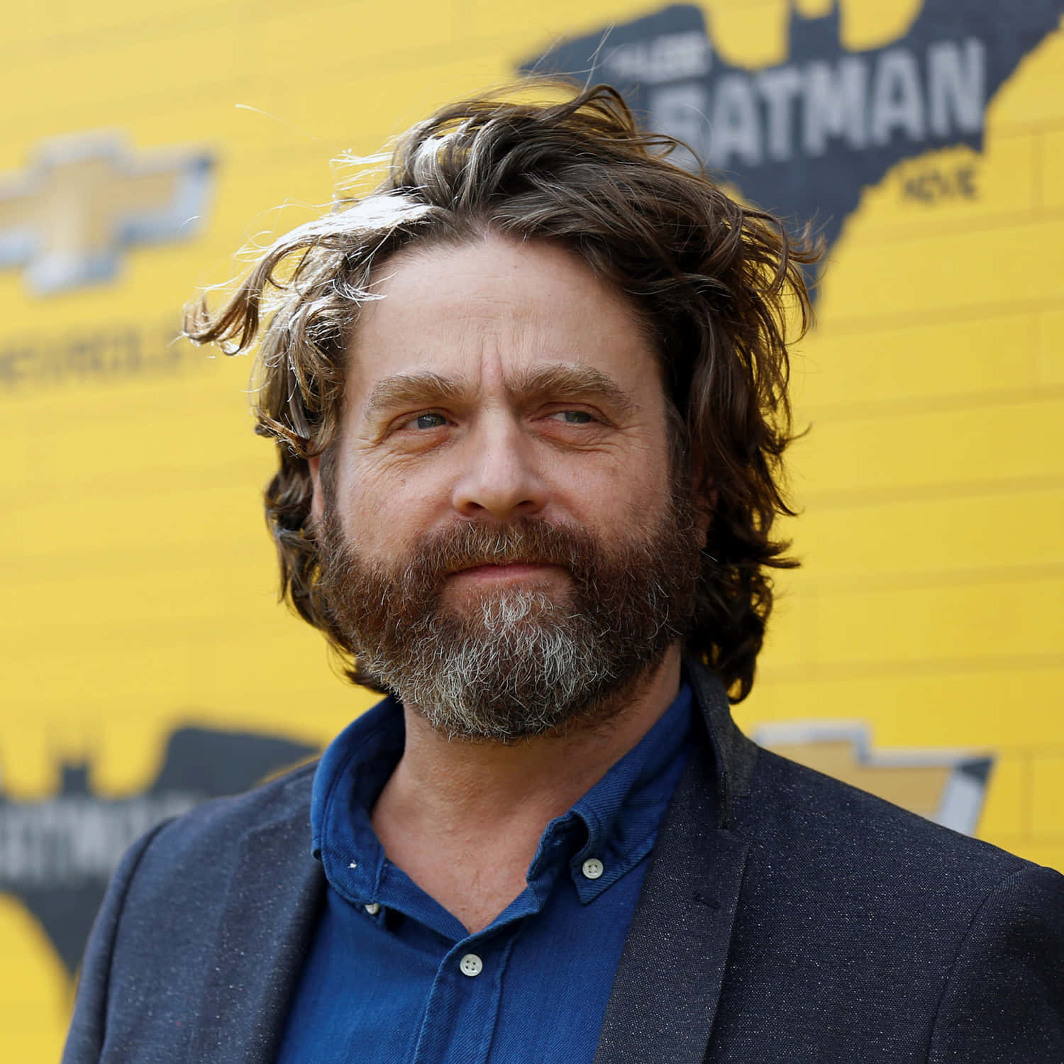 Zach Galifianakis 桌布 壁紙 — 一位留著灰白鬍鬚、頭髮凌亂的男子，在明亮的黃色蝙蝠俠電影背景前露出微妙笑容。