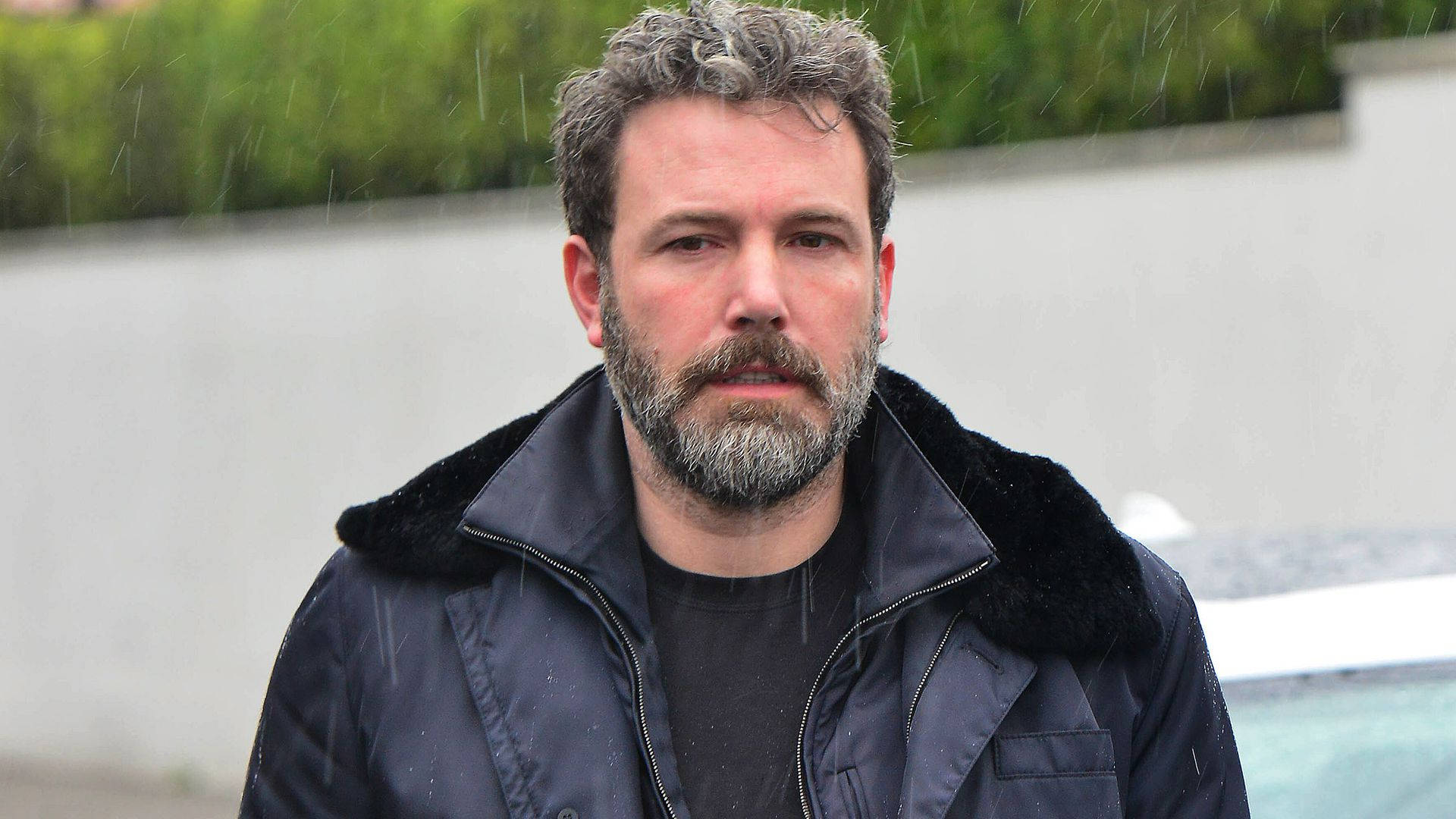 Ben Affleck 桌布 壁紙 — 一位留著灰白鬍鬚和捲髮的男子，穿著帶毛領的深色外套站在雨中。