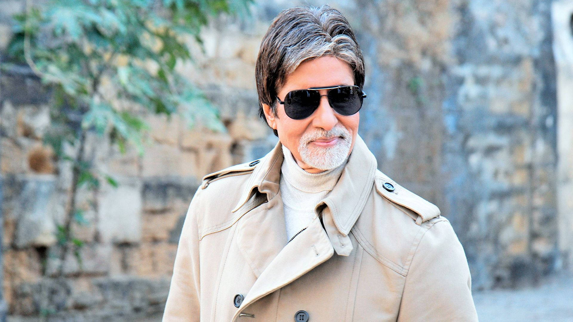 Amitabh Bachchan 桌布 壁紙 — 一位留著灰色鬍鬚、戴著太陽眼鏡的時尚男士，身穿米色風衣，背景是石牆。