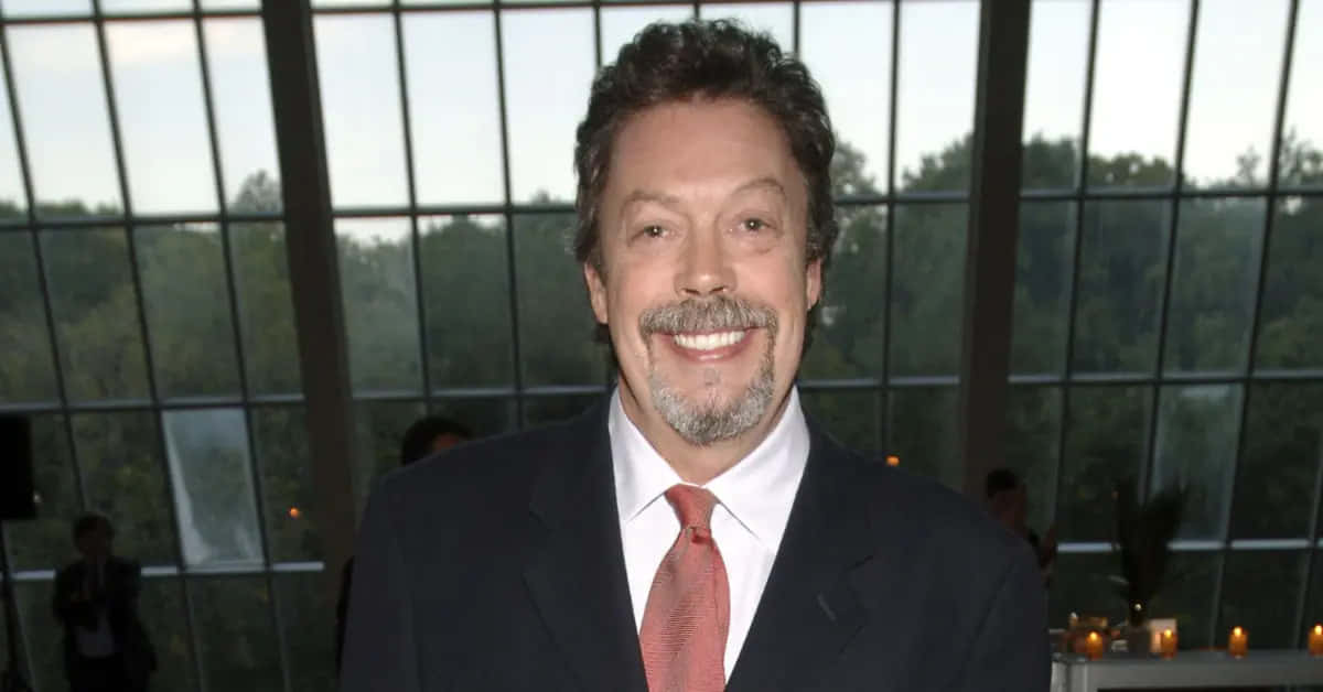 Tim Curry 桌布 壁紙 — 一位留著灰色鬍鬚和鬍子的微笑男士，身穿深色西裝和紅色領帶，背景是大窗戶，窗外是綠樹。