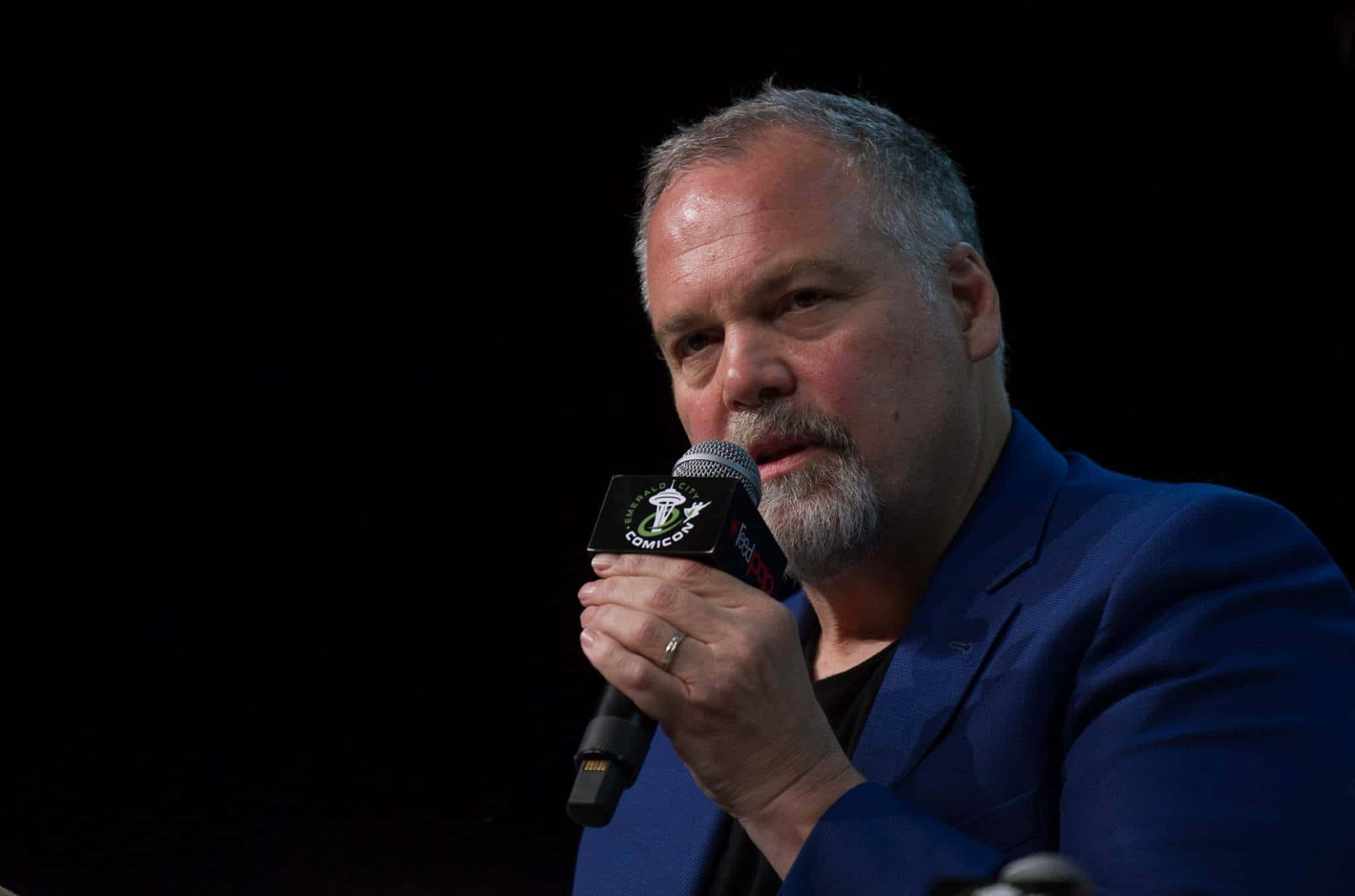 Vincent D'onofrio 桌布 壁紙 — 一位留著灰色鬍鬚的男子在黑色背景的漫畫展上對著麥克風說話。